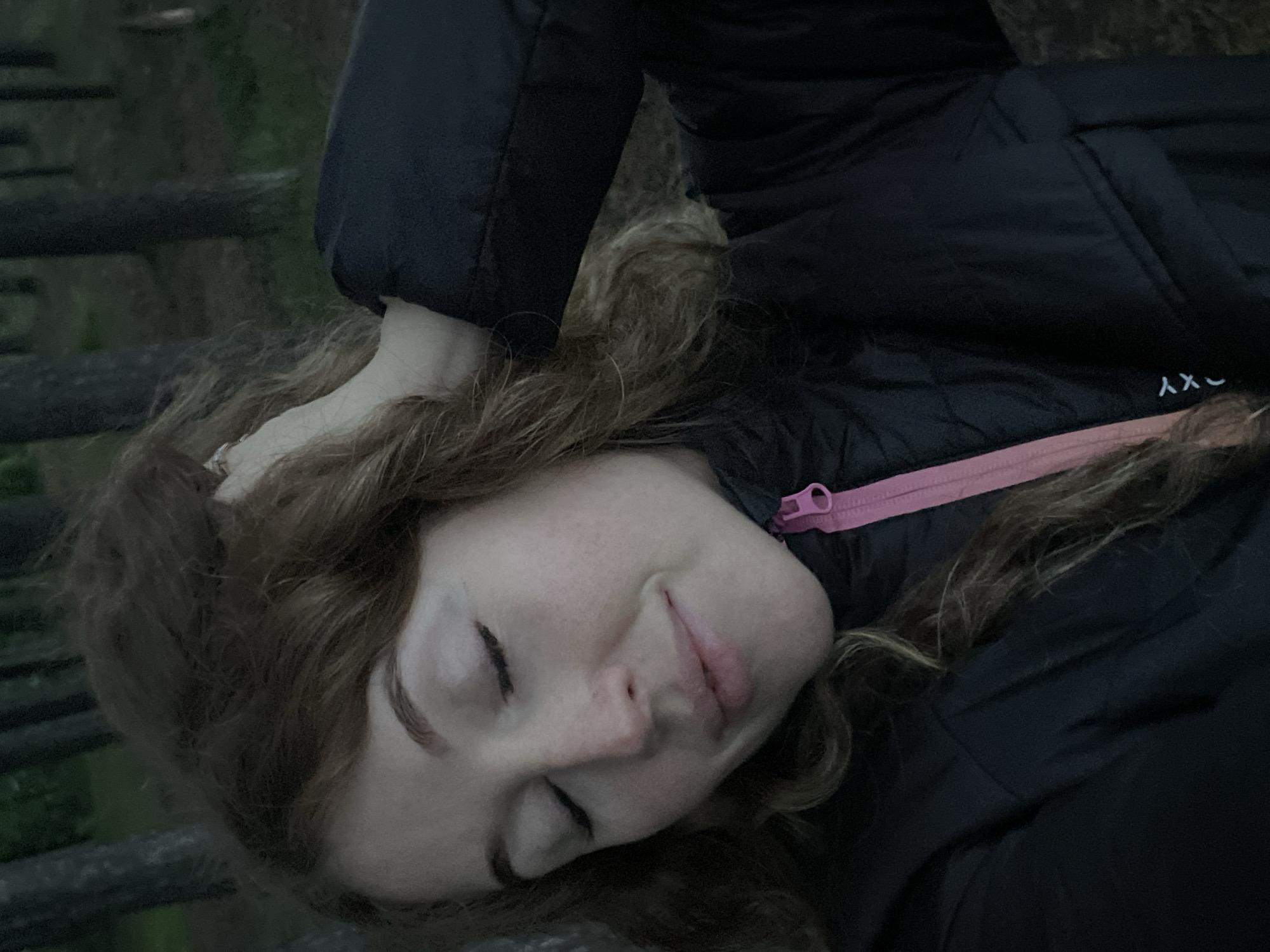 A woman is lying down outdoors. She has fair skin, long brown curly hair, and is wearing a black puffy jacket with a pink zipper. Her eyes are closed and she has a slight smile. She is lying on what appears to be a wooden structure or platform. The location is unknown.