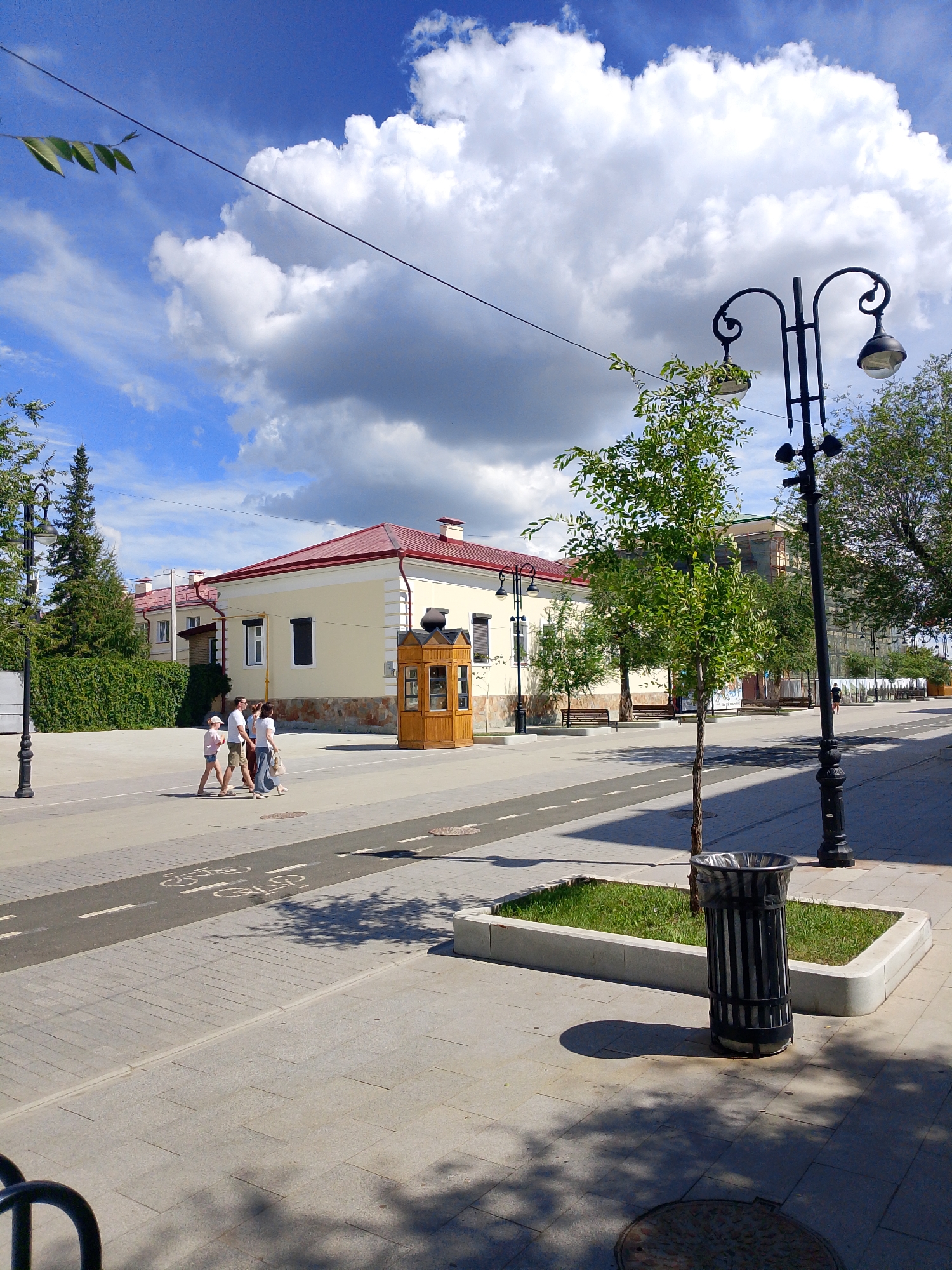 A street scene in Orenburg, Russia, features a paved walkway with a designated bicycle lane marked with white paint. A black trash receptacle stands on a small patch of grass enclosed by a concrete border. In the middle ground, four people are walking across the street. To the left of the people, a small wooden kiosk or booth is situated on the walkway. A building with a red roof and cream-colored walls is visible behind the kiosk and the walking individuals. The sky is a vibrant blue, populated with large, white, fluffy clouds. A black lamppost with two lights stands on the right side of the frame, and another lamppost is visible on the far left. Trees line the street, with some providing shade on the walkway, casting dappled shadows. The overall impression is a bright, sunny day in an urban environment.