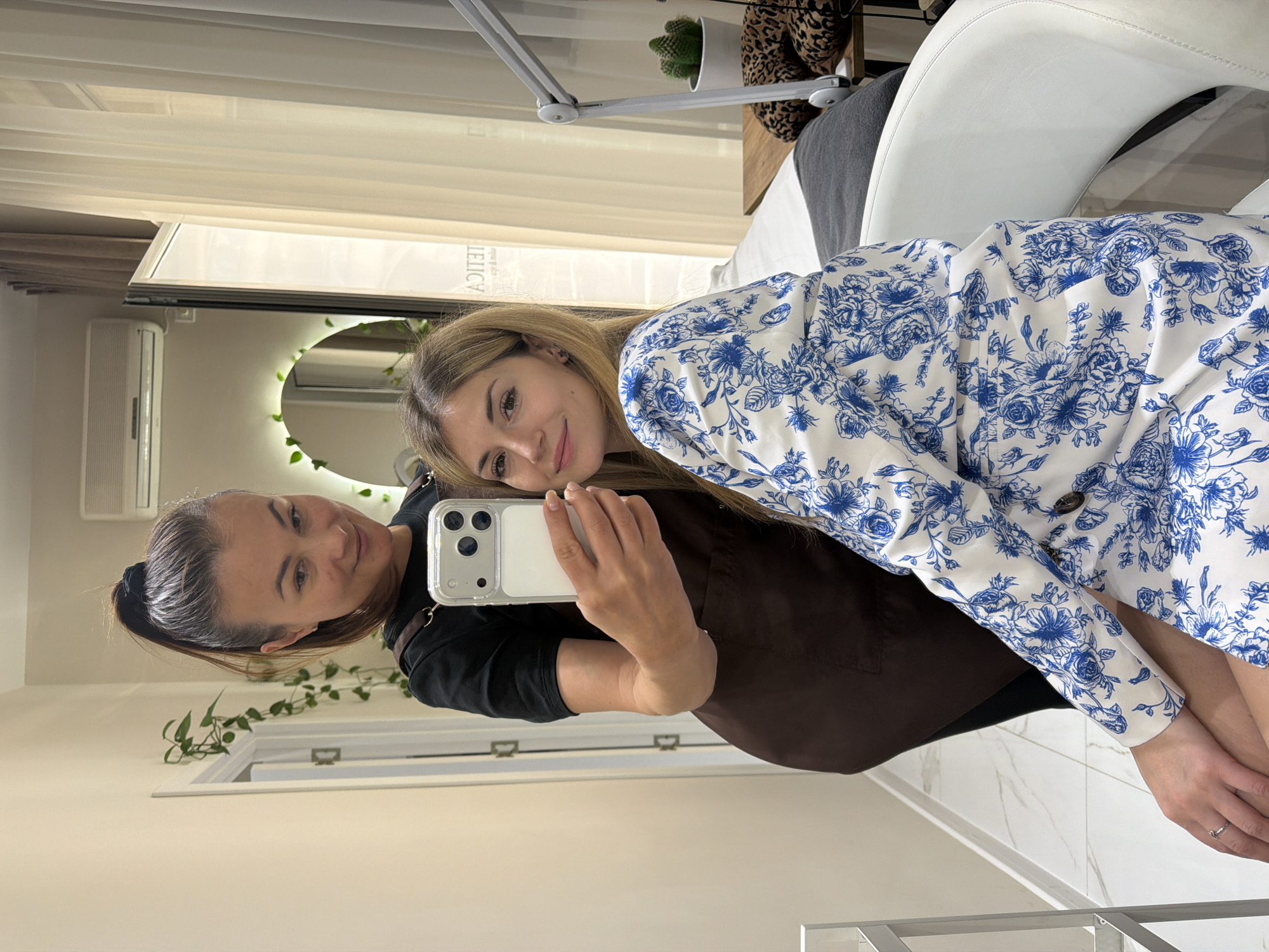 The image shows two women taking a selfie. The woman on the left is holding a smartphone in her right hand and has her left arm around the other woman, who is on the right side of the image. She has dark hair pulled back in a ponytail and is wearing a black t-shirt and a brown apron. The woman on the right has blonde hair and is wearing a white jacket with a blue floral pattern. The women are indoors, in a room with a white and light beige color scheme. In the background, there is a round mirror with lights and greenery, an air conditioning unit, and white curtains. There is a white chair and a desk with a lamp and decorative items. Based on the interior design and context clues, the image may be taken in a beauty salon or spa in Budva, Montenegro. There is no definitive visual confirmation of the exact location.