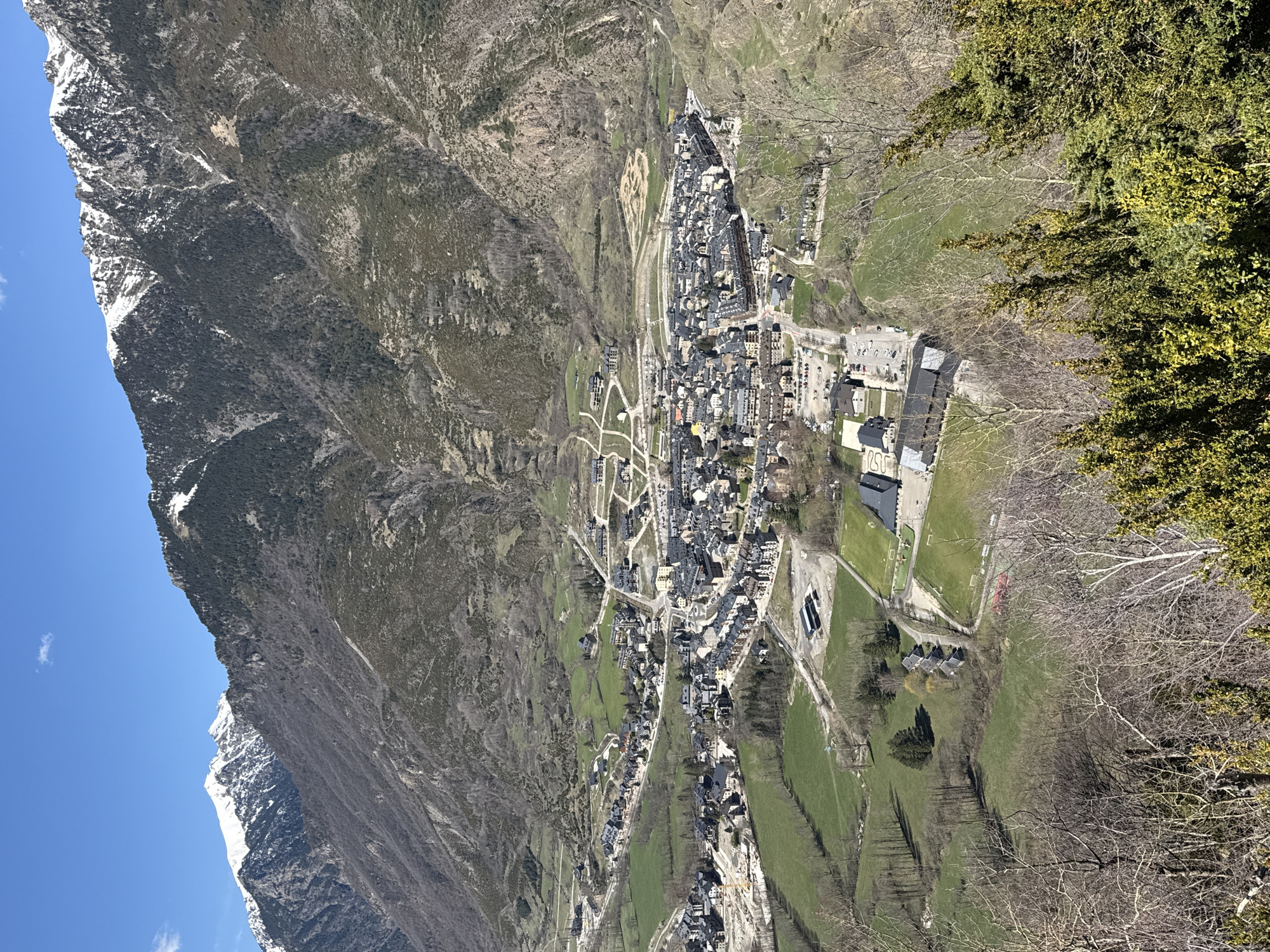 The image presents an aerial view of the city of Benasque, Spain, nestled in a valley between mountains. The foreground includes the rough terrain of the valley slopes with trees and sparse vegetation.
The town of Benasque is densely packed with buildings, primarily structures with dark roofs, arranged in a network of streets. To the east of the town are grassy fields with some sparse trees and sports facilities.
The background features a snow-capped mountain range, part of the Pyrenees. The sky visible to the left is blue, suggesting clear weather conditions.