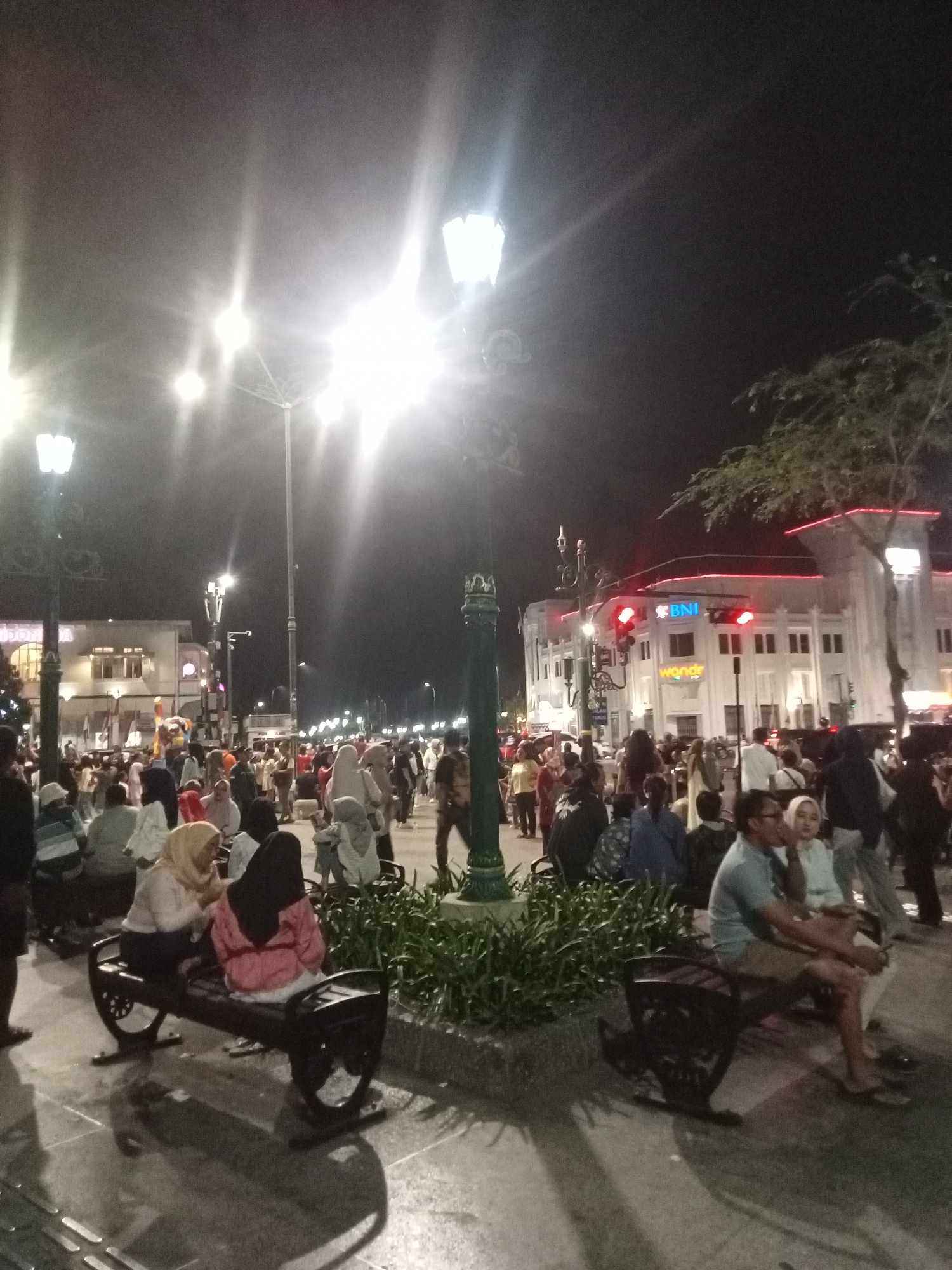 This image depicts a nighttime scene in the city of Yogyakarta, Indonesia, likely at Malioboro Street, a popular pedestrian area known for its vibrant atmosphere. A crowd of people, dressed in casual attire, are seen walking and gathering. Some individuals are seated on benches, observing the surroundings or interacting with each other. Streetlights illuminate the scene, casting long shadows on the pavement. In the background, buildings with illuminated windows and signage are visible, including one with a prominent "BNI" logo, suggesting a financial institution. The overall impression is that of a lively urban environment with a significant number of people enjoying the evening.