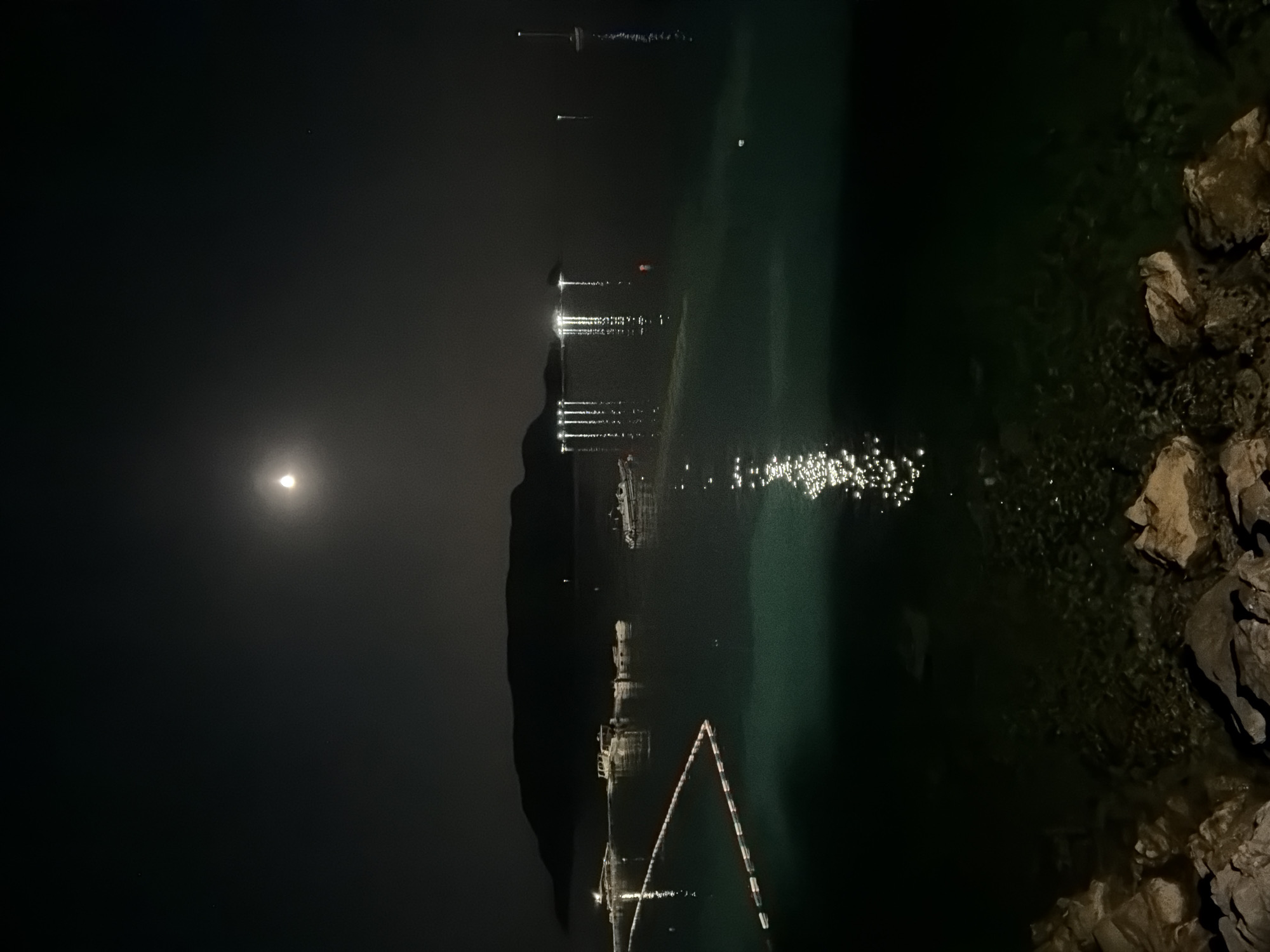 The image depicts a nighttime scene of a bay in Budva, Montenegro. The moon is visible in the dark sky, casting a faint glow and creating reflections on the calm, dark green water. Several boats are anchored or moored in the bay, some of which are illuminated by lights. A buouy system, forming a triangular shape, is visible in the foreground, marking an area in the water. The shoreline in the foreground consists of rocks and appears to be a waterfront promenade or beach. In the background, a dark, silhouetted island or peninsula rises from the sea. The overall atmosphere is tranquil and serene, with the lights from the boats and the moon contributing to the nocturnal ambiance.