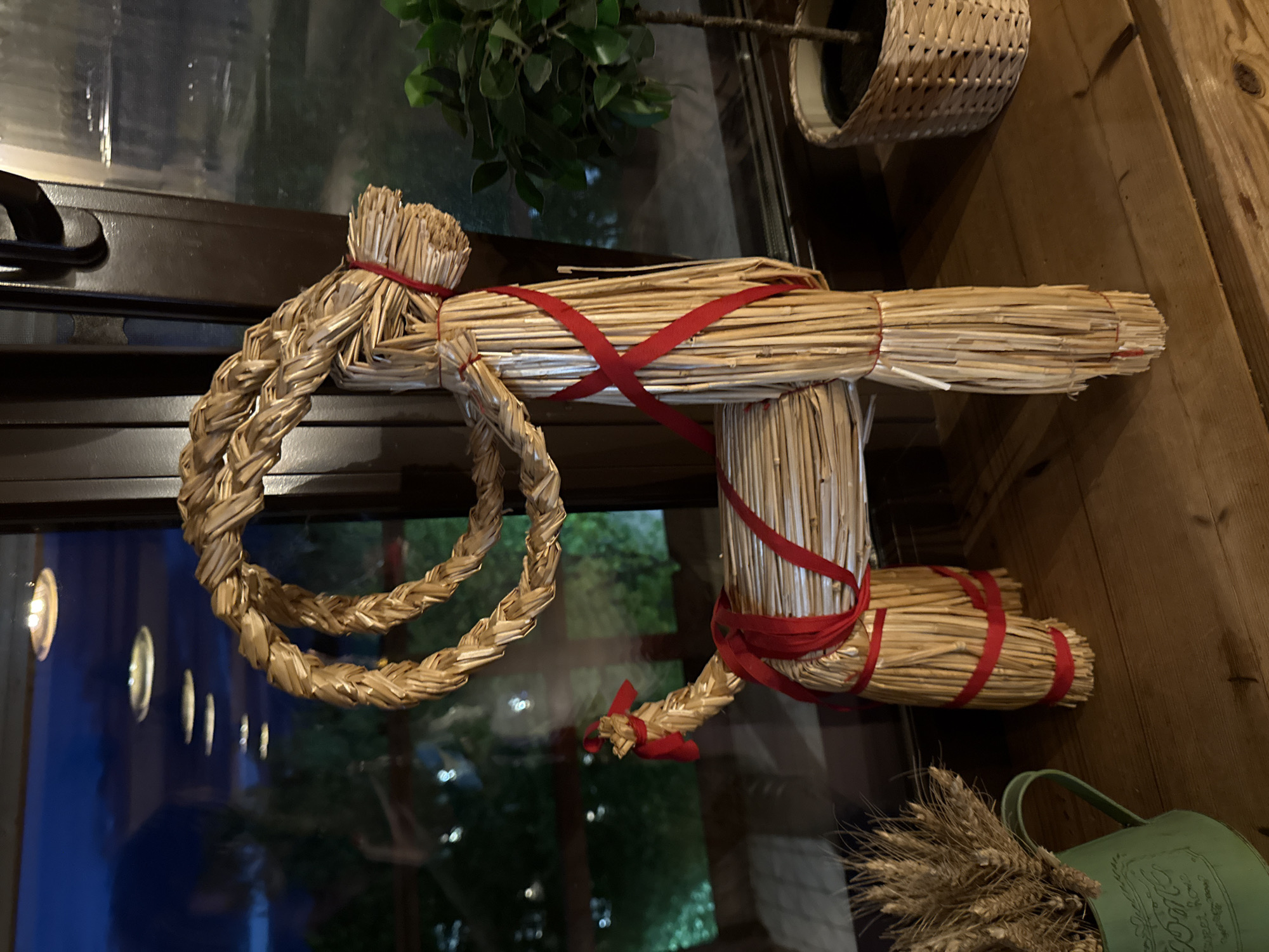 An indoor scene depicts a straw doll figure tied with red ribbons, positioned near a window. The figure has a circular braided element forming part of its design. The scene includes elements such as plants, a wicker basket, and a decorative watering can. The specific location is the city of Peresvet, Russia.