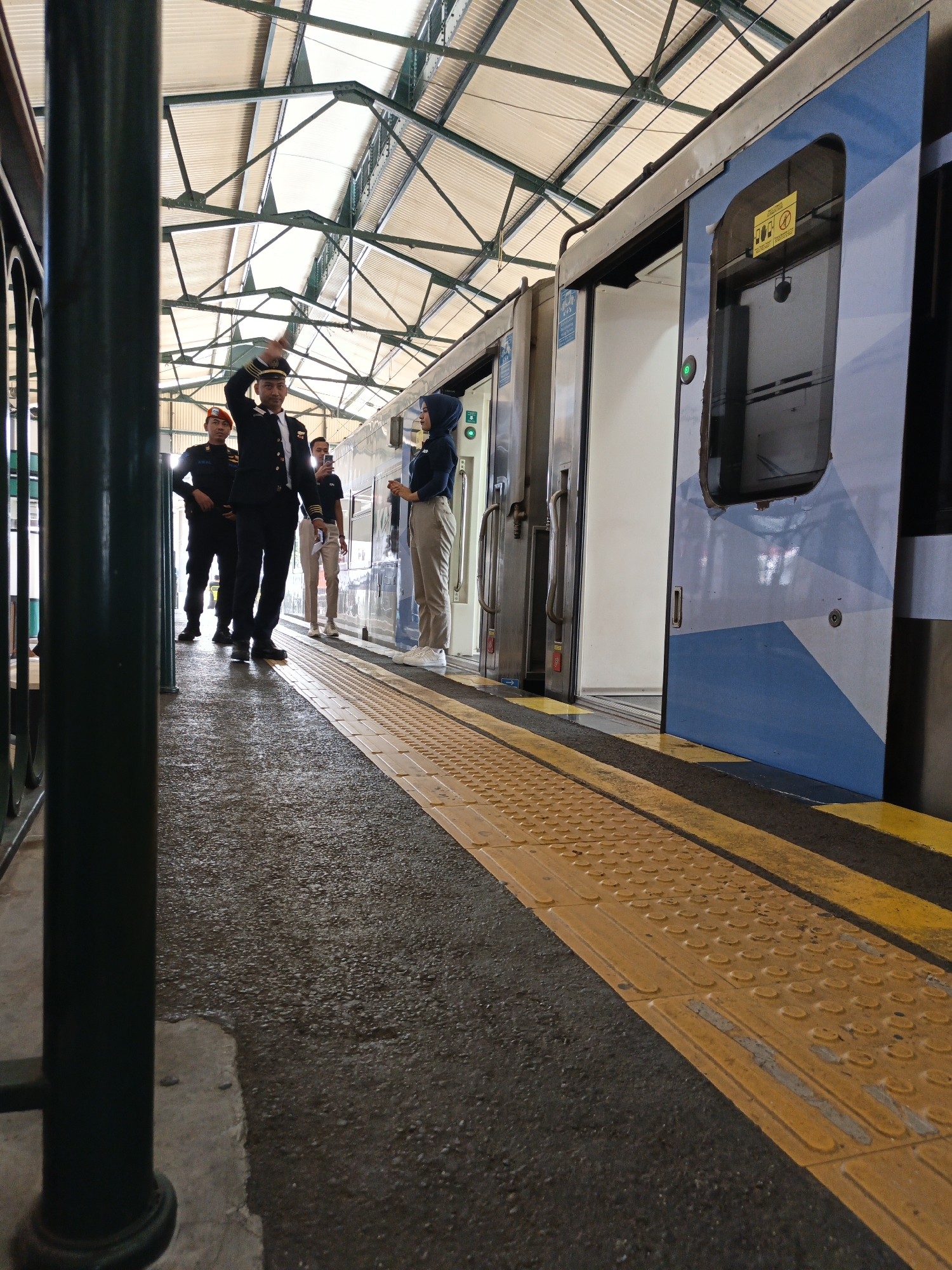 A low-angle, medium shot captures a train platform in Klaten, Indonesia, under a covered roof with a visible skeletal support structure. The train, partially open with its doors ajar, stands adjacent to the platform. A series of yellow tactile paving tiles are prominently featured on the platform edge, indicating a safety feature for visually impaired individuals.
Several individuals are present on the platform. A man in a dark uniform, adorned with medals and a cap, stands in the foreground, holding his cap aloft as if in greeting or salute. To his left, another person in a dark uniform and an orange helmet is visible. Further back, a person in a light-colored shirt and pants holds a smartphone up, possibly taking a photo. On the right side of the train, a woman wearing a hijab and a dark shirt over light-colored pants stands near the train door.
The ground of the platform appears to be asphalt, with some wet patches suggesting recent rain or cleaning. The overall atmosphere is that of a train station during a service or inspection.