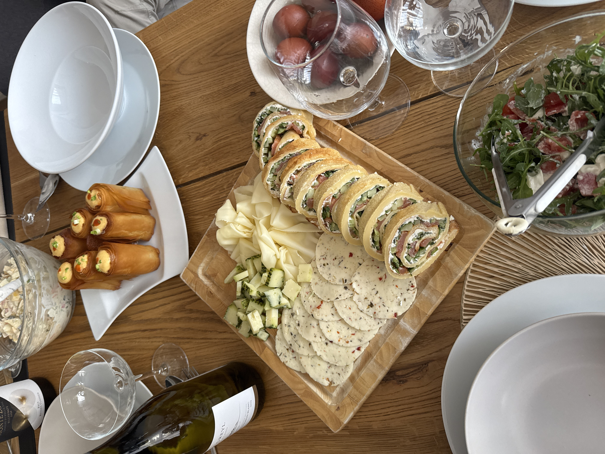 The image shows a wooden table laden with food and tableware, suggesting a meal or gathering. A wooden serving board holds rolled crepe-like appetizers, various cheeses (sliced, cubed, and seasoned), and crackers. Nearby are dishes containing salad with greens and tomatoes, fried spring rolls, and a potato salad. White ceramic bowls and plates are stacked at the edges of the table. Wine glasses and a bottle of white wine are also visible. The scene appears to be indoors, likely in a home or restaurant setting. The location cannot be determined from the image, and the assumption of being in Warsaw, Poland is not supported by visual information.