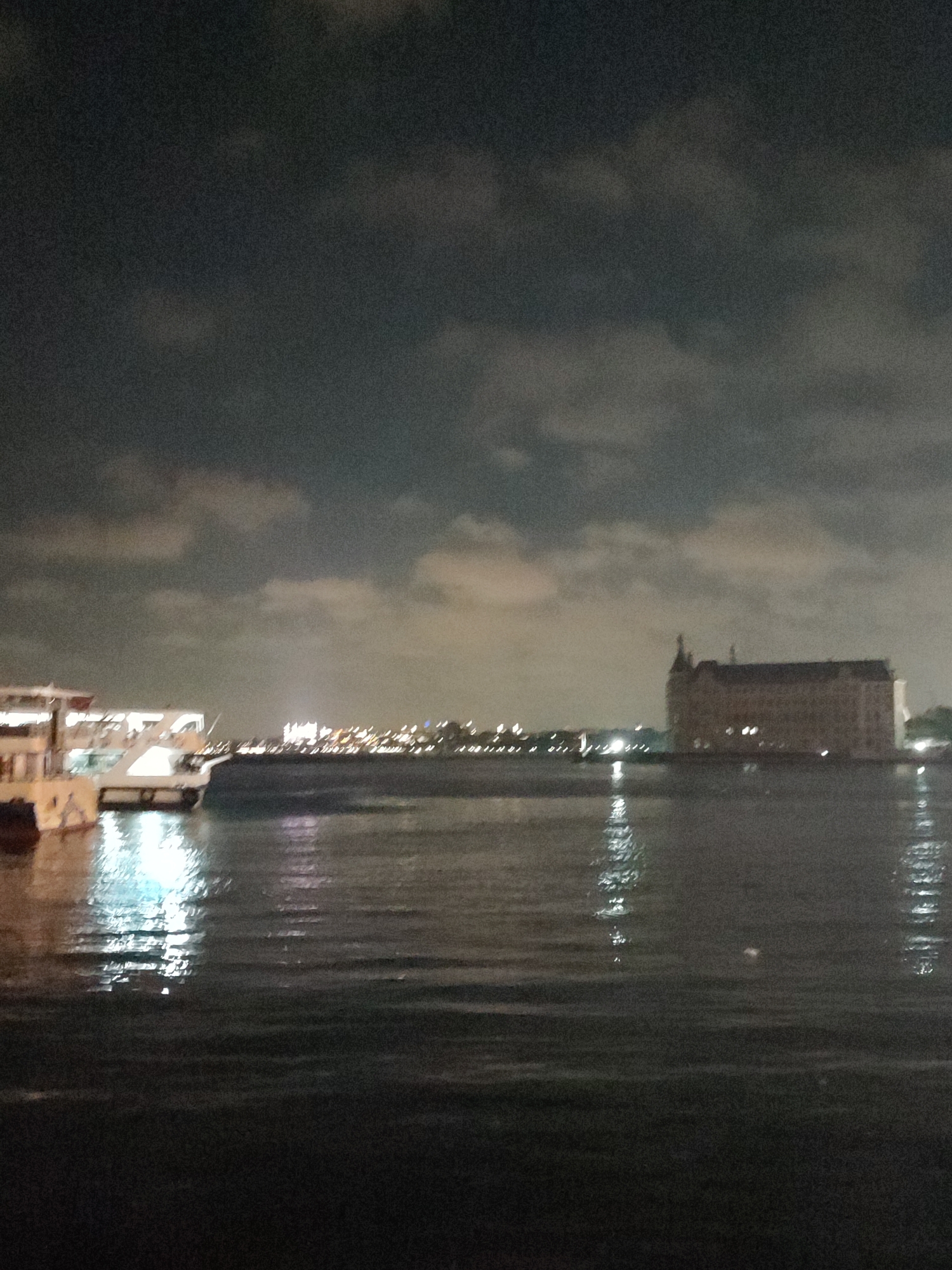 The image depicts a nighttime view of the waterfront in Kadıköy, Turkey. On the left side, a large, illuminated ferry is docked, its bright lights reflecting on the dark water. In the distance, across the water, the skyline of another part of the city is visible with numerous lights. Prominently featured on the right side of the image is the Haydarpaşa Train Station, a large, historic building with illuminated windows, standing against the dark sky. The water is calm, with gentle ripples reflecting the lights from the shore and the ferry. The sky is cloudy and dark, suggesting it is late at night or early morning. The overall scene conveys a sense of urban activity and architectural grandeur at night.
