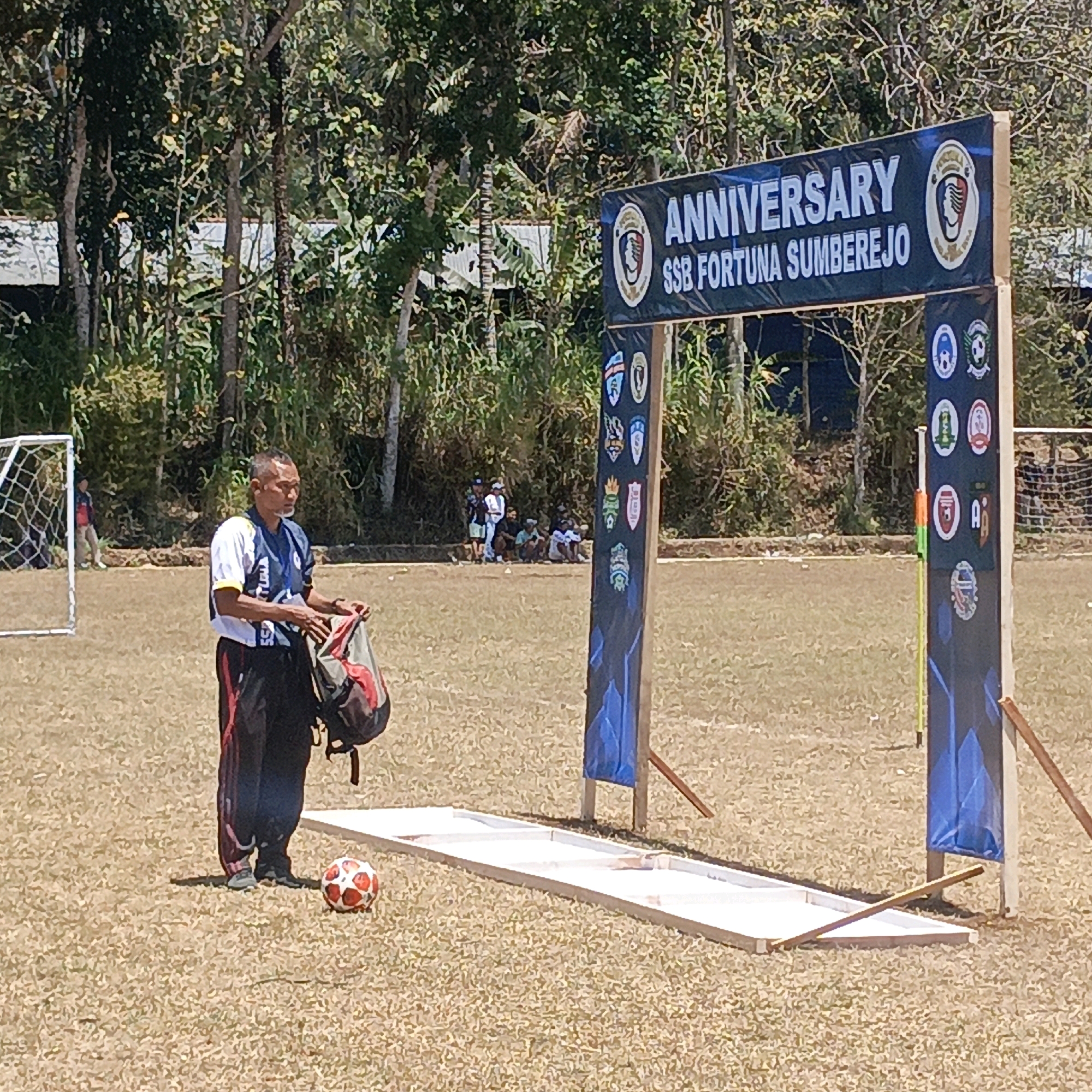 In Krajan Lor, Indonesia, a man stands on a dry, grassy soccer field, holding a red and grey backpack. He is wearing a white shirt with black and red accents and black pants. In front of him, a soccer ball rests near a white, wooden structure. Behind the man, a banner hangs over an archway that reads "ANNIVERSARY SSB FORTUNA SUMBEREJO". The archway is decorated with several emblems and logos. In the background, trees line the edge of the field, and a soccer goal is visible to the left. Several people are seen sitting or standing in the background near the trees.