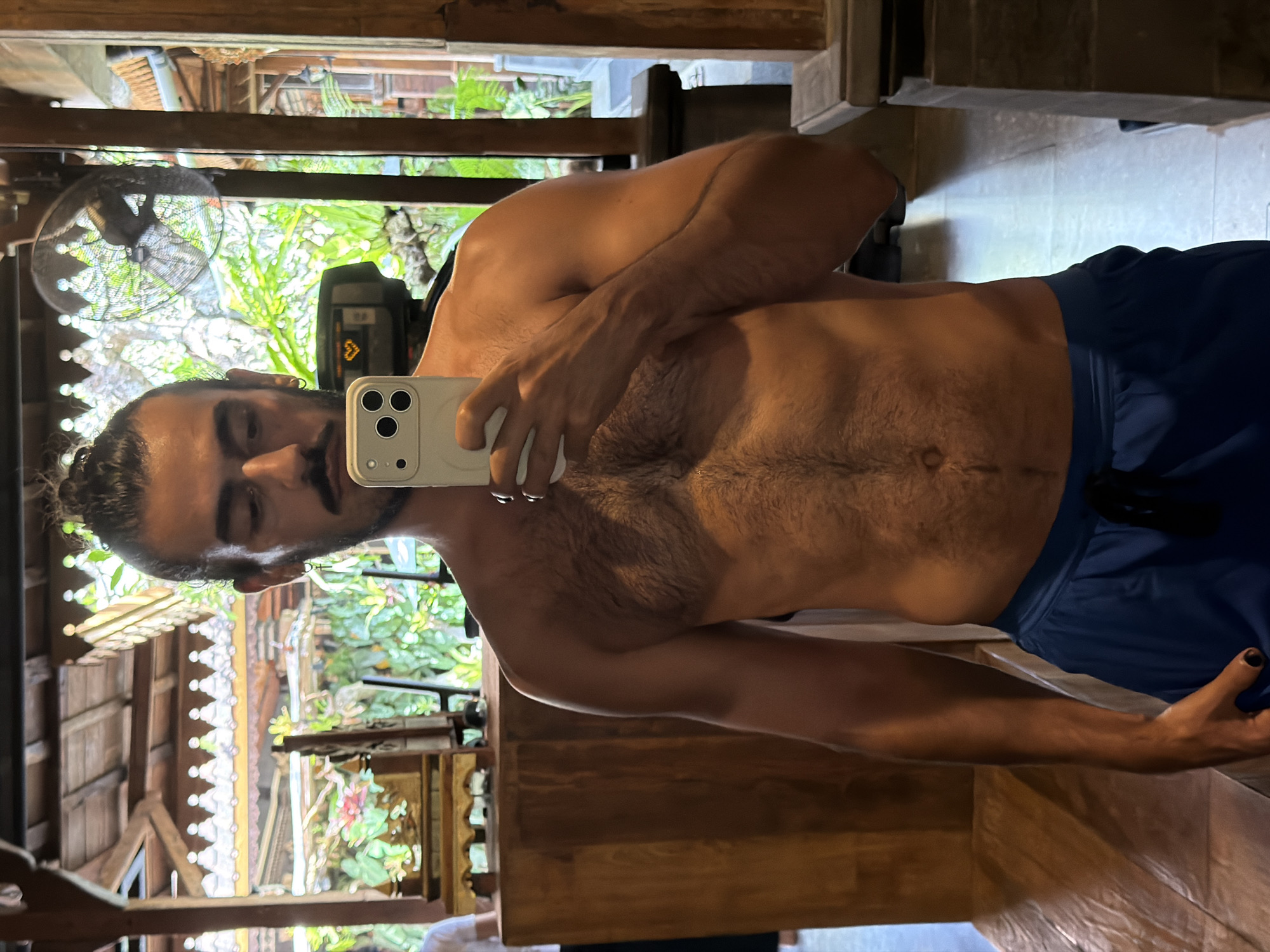 A shirtless man with a bun and mustache stands in front of a mirror, taking a selfie. He is wearing blue athletic shorts. The background appears to be a resort or hotel in North Kuta, Indonesia, with intricate wooden architecture and lush greenery visible through a fan. A piece of exercise equipment is reflected in the mirror behind him.