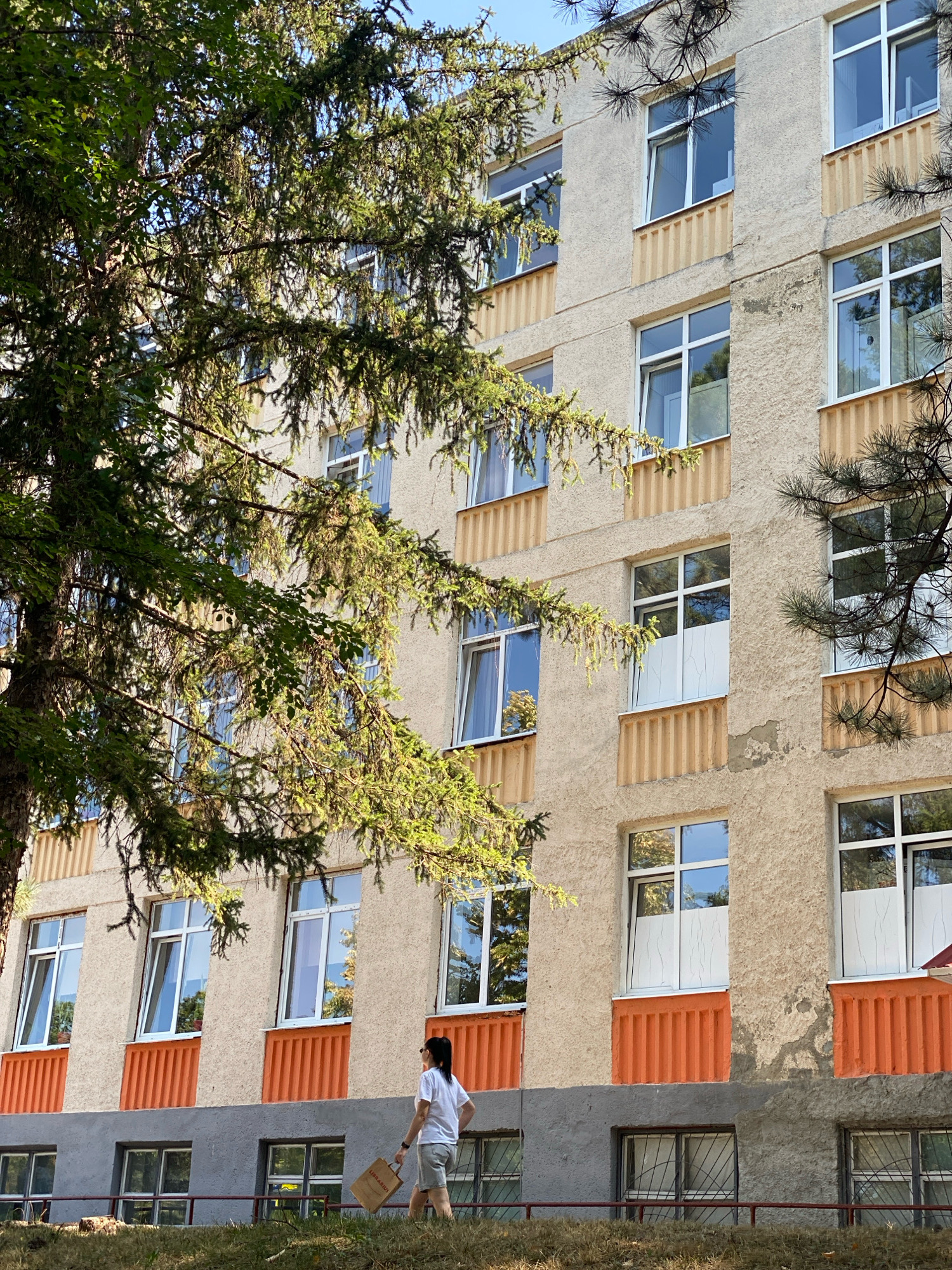 An outdoor, low-angle shot captures the side of a multi-story building with a facade of beige stucco and orange vertical accents below the windows. Pine branches overhang from the left, casting dappled sunlight onto the building and a person walking from right to left in the foreground. The person is a woman with dark hair pulled back, wearing a white t-shirt and light-colored shorts. She is carrying a brown paper bag in her right hand. Below the woman, a grey-colored base level of the building is visible, with barred windows. The scene appears to be in Chișinău, Moldova, based on the architectural style and common building types found there.