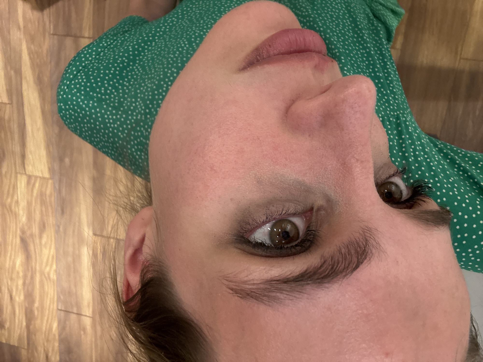 A close-up, angled view of a person's face. The person has light skin, brown eyes, and dark eyebrows. The person's upper body is clad in a green dress with white polka dots. The background appears to be a light-colored wooden surface. The setting is Unknown, Unknown.