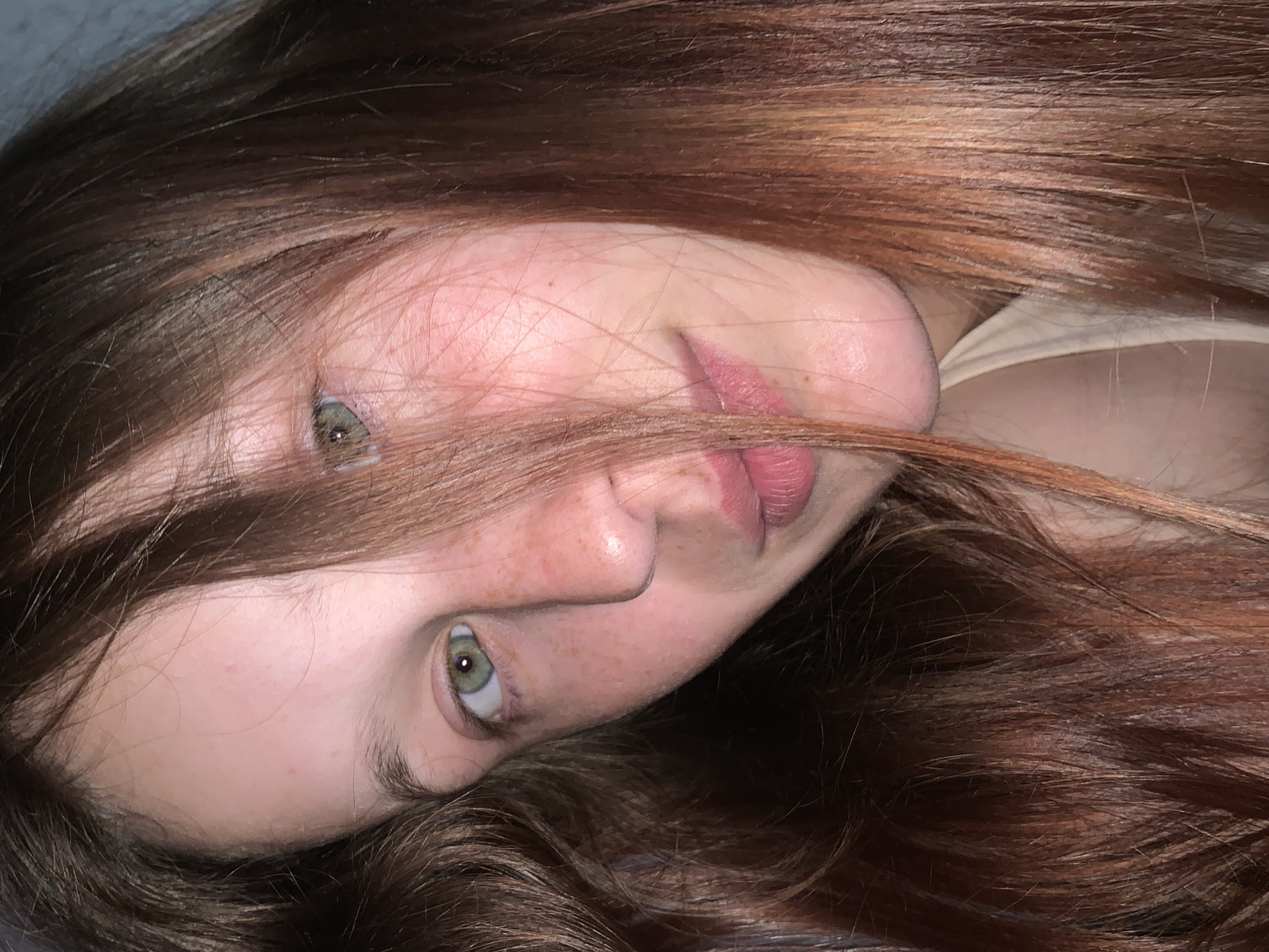 This close-up portrait features a young individual, likely female, with fair skin, prominent reddish-brown hair, and green eyes. The subject's head is tilted significantly to the side, or she is lying down, with her voluminous hair fanned out, creating a textured backdrop. Strands of hair partially cover her face, notably obscuring parts of her forehead and one eye. Her visible eye, a striking green, looks directly towards the viewer, while the other is partially hidden. Her nose has a natural contour, and her lips, soft and pink, are slightly parted in a relaxed manner, not quite smiling. Small freckles or natural skin variations are visible across her cheeks and nose. A light-colored garment, possibly a shoulder strap or part of a top, is visible on the right side of the frame, near her shoulder. The lighting is soft and even, suggesting an indoor setting or diffused natural light. The overall mood is calm and contemplative. No specific activity is shown beyond posing for the photograph. There is no visible text. The photograph is set in Foki, Russia.