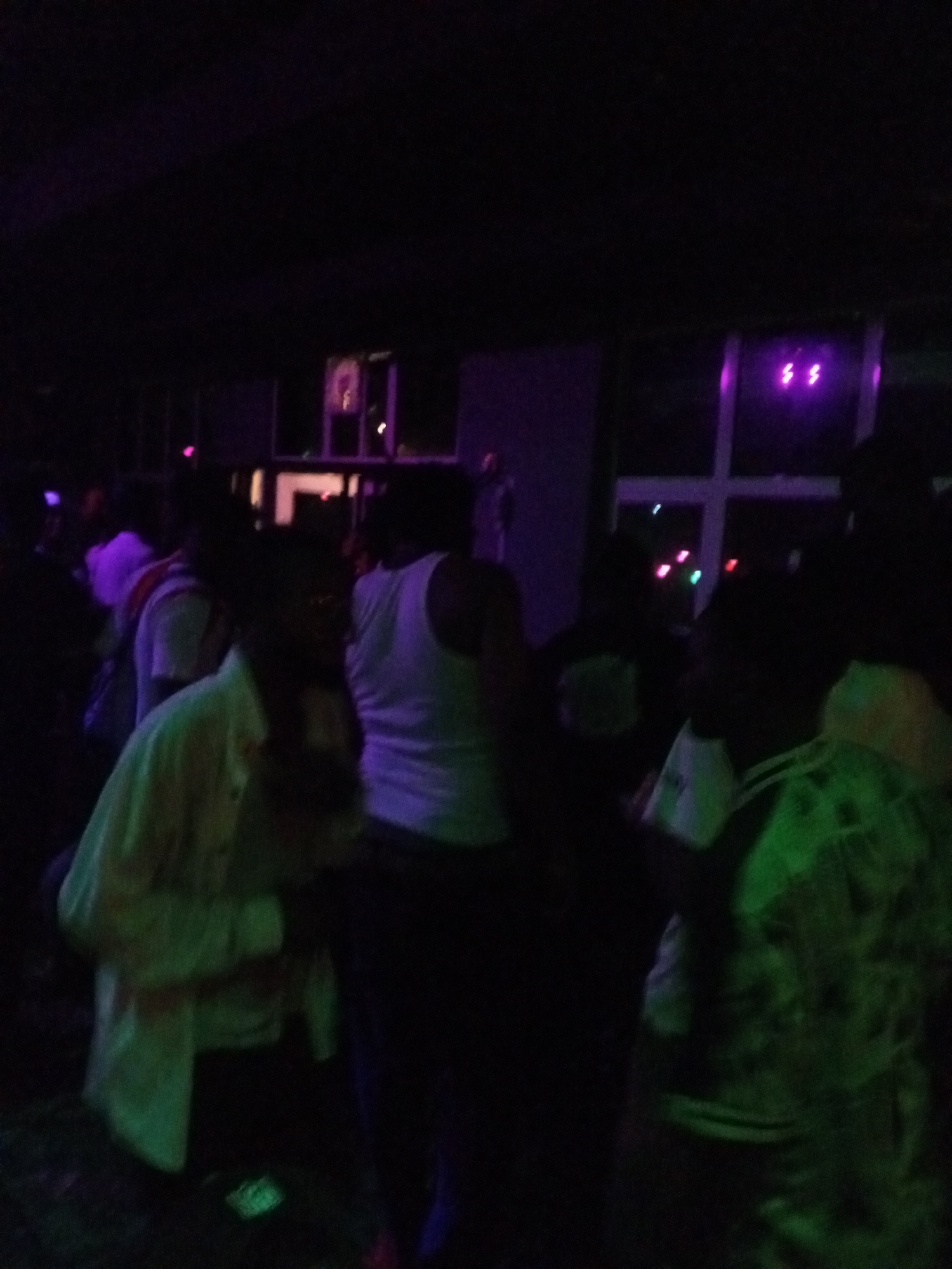 The image shows a dimly lit indoor setting, possibly a nightclub or party venue in Lokoja, Nigeria. Multiple individuals are present, primarily facing away from the camera. A person in the center wears a white tank top. Another individual on the left wears a light-colored shirt. The environment is dark with indistinct lighting. The background features a window or opening. Due to the low light, details are obscured. The location in Lokoja cannot be determined specifically.