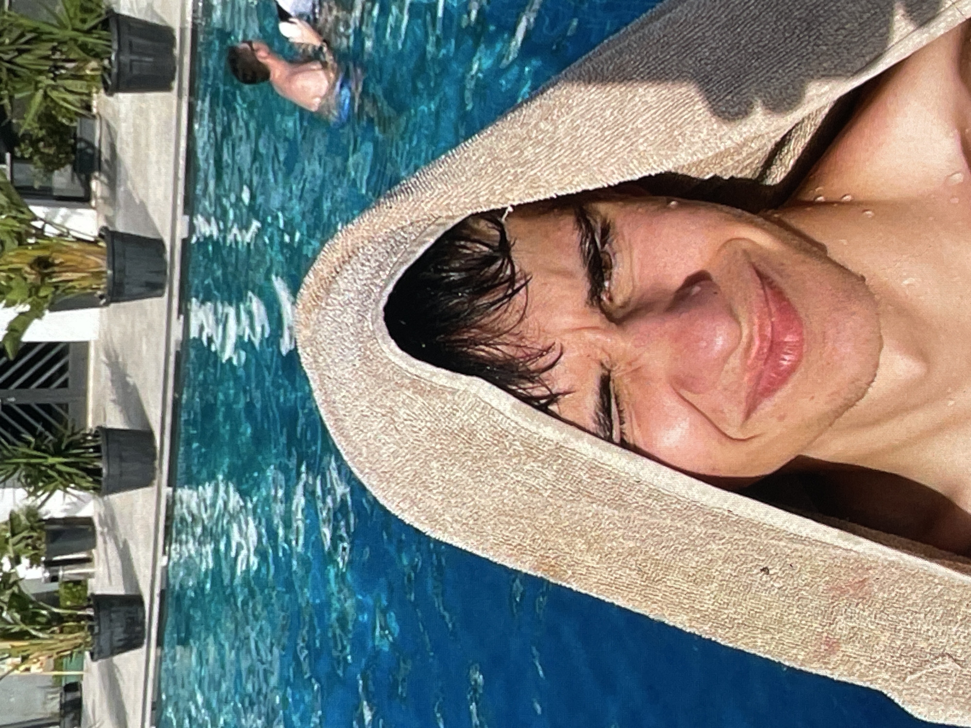 The image shows a close-up selfie of a young man with a towel draped over his head, taken in a swimming pool. He is partially submerged in the blue water, with his face turned towards the camera, a wide smile, and his eyes squinting. Droplets of water are visible on his skin and hair. In the background, out of focus, are two other individuals in the pool and potted plants on the poolside. The scene appears to be outdoors, possibly at a resort or private residence. The location of Harmanli, Bulgaria is not indicated in the image.