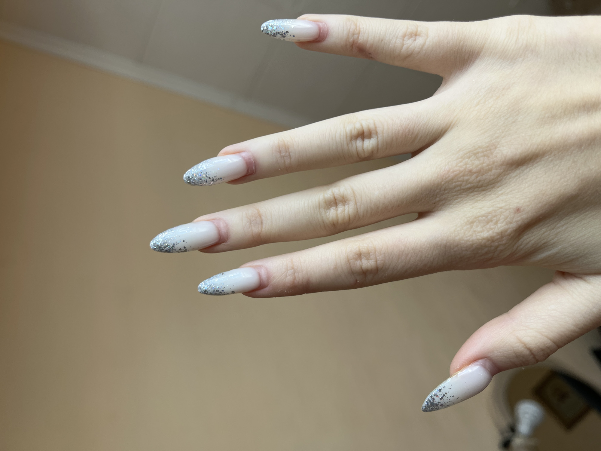 A close-up, medium shot captures a person's left hand with light skin and long, almond-shaped nails. The nails are painted with a pale, milky white polish, and the tips are adorned with a gradient of silver glitter. The hand is positioned in the foreground, with the fingers gently curved and slightly spread. The background is a soft-focus, neutral beige wall with a subtle horizontal line near the top, indicative of a room interior. There is no visible indication of the city of Ivanovo, Russia. The lighting is soft and diffused, highlighting the texture of the skin and the sparkle of the glitter. The overall impression is one of a person showcasing their freshly manicured nails.