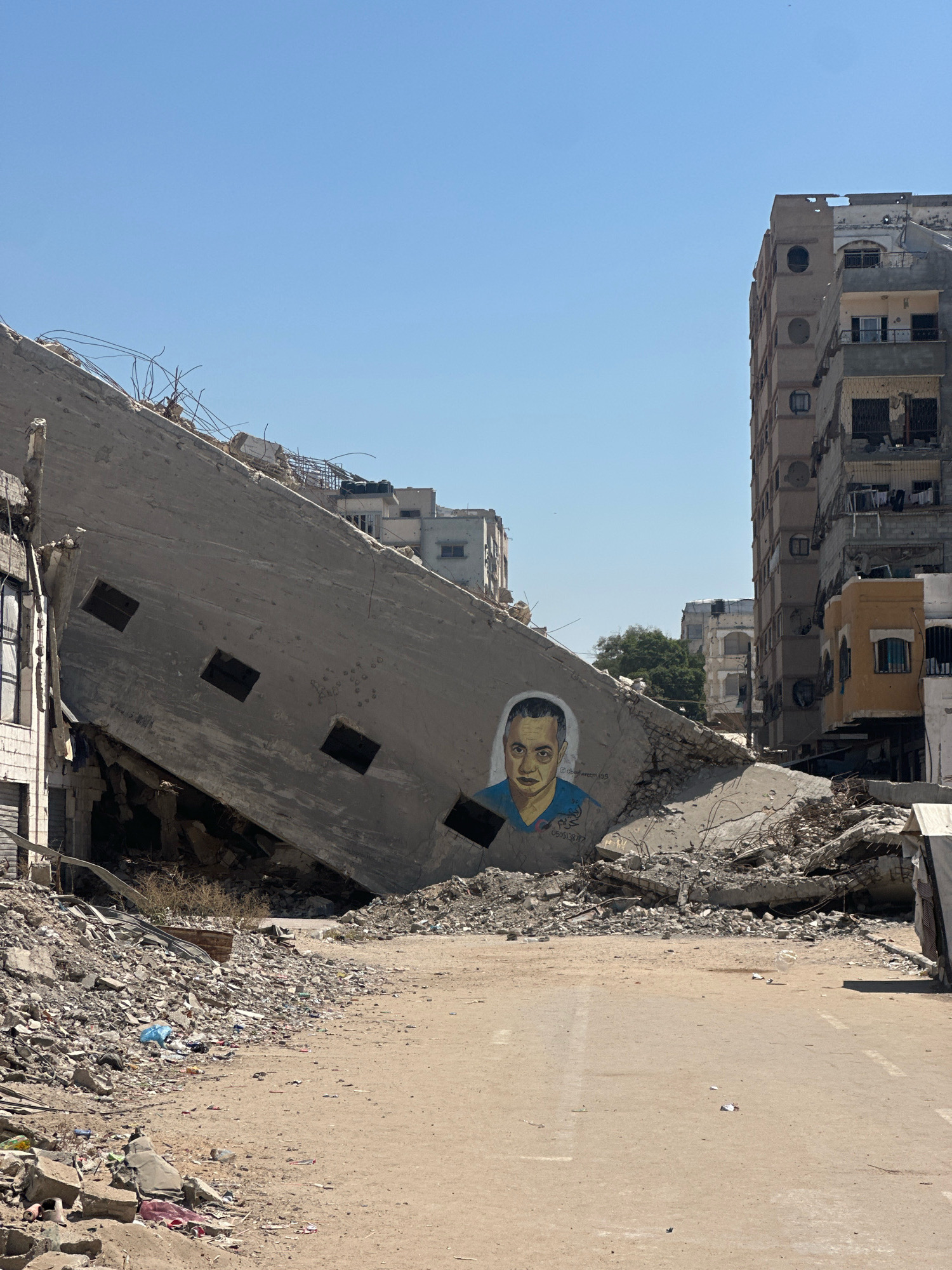 The image depicts a scene of destruction in the city of Gaza, Palestinian Territories, under a clear blue sky. In the foreground, a large, angled concrete slab, part of a demolished building, leans precariously. A mural of a man's face, painted in blues and yellows, is visible on this slab. The man has short dark hair, dark eyes, and a slight frown. Below the mural, there is a phone number written in black. The ground around the slab is covered in rubble, including concrete fragments, twisted rebar, and scattered debris. In the background, damaged multi-story buildings stand, some appearing partially intact while others show signs of significant structural damage. Clothes hang from balconies on one of the intact buildings. The overall environment suggests the aftermath of conflict or extensive destruction.