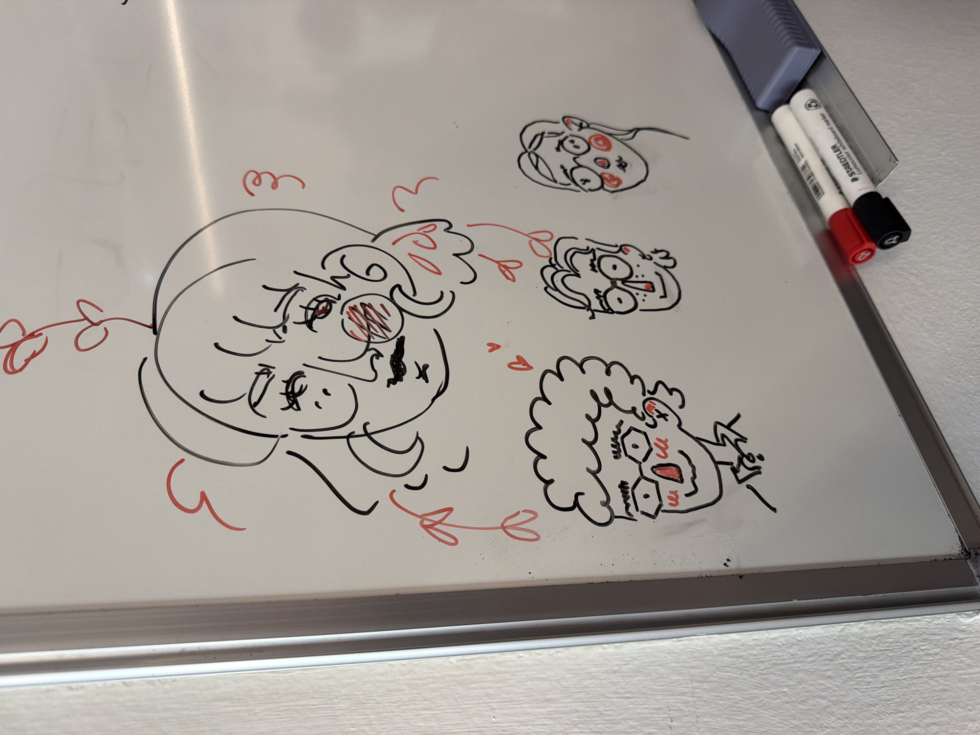 The image depicts a whiteboard with cartoonish drawings in black and red markers. The drawings appear to be caricatures of faces with exaggerated features. Two markers (red and black) are visible next to the whiteboard. The image lacks contextual information that would link it to Milan, Italy.