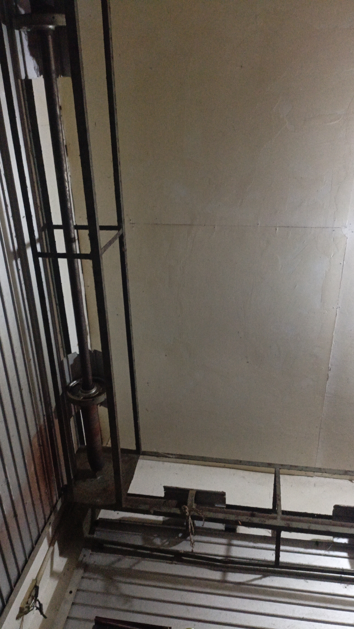 A metal frame structure is visible in this interior scene, likely within a building. On the left side of the image, vertical metal bars form a partial enclosure, with a rusty pipe running vertically within the frame. This pipe appears to be attached to some sort of circular mechanism near its base. The floor area under this structure is dimly lit, with evidence of horizontal metal supports and some white rectangular panels. The background consists of a plain, light-colored wall, possibly plaster, with some subtle texture and discoloration. The lighting suggests an indoor environment, possibly with a single light source illuminating the upper part of the wall. There are no people or animals present in the image. The overall impression is one of a utilitarian or unfinished space.