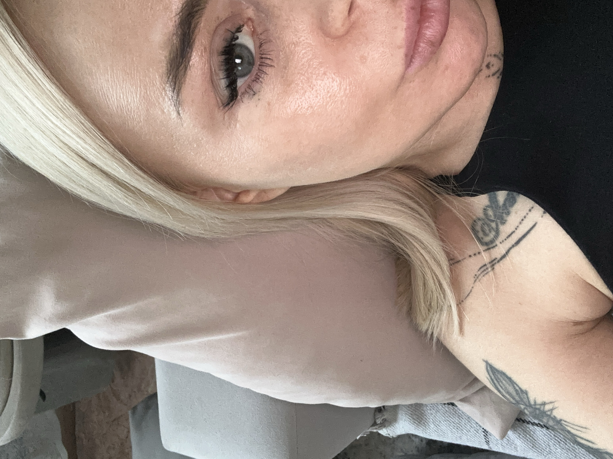A close-up, angled portrait of a light-skinned person with blonde hair. The person has light eyes, full lips, and visible tattoos on their arm and near their ear. The person is wearing a neutral-colored shirt, and the background includes a dark-colored garment and a neutral-toned fabric. The image location, Высокий, Russia, cannot be verified from the content of the image.