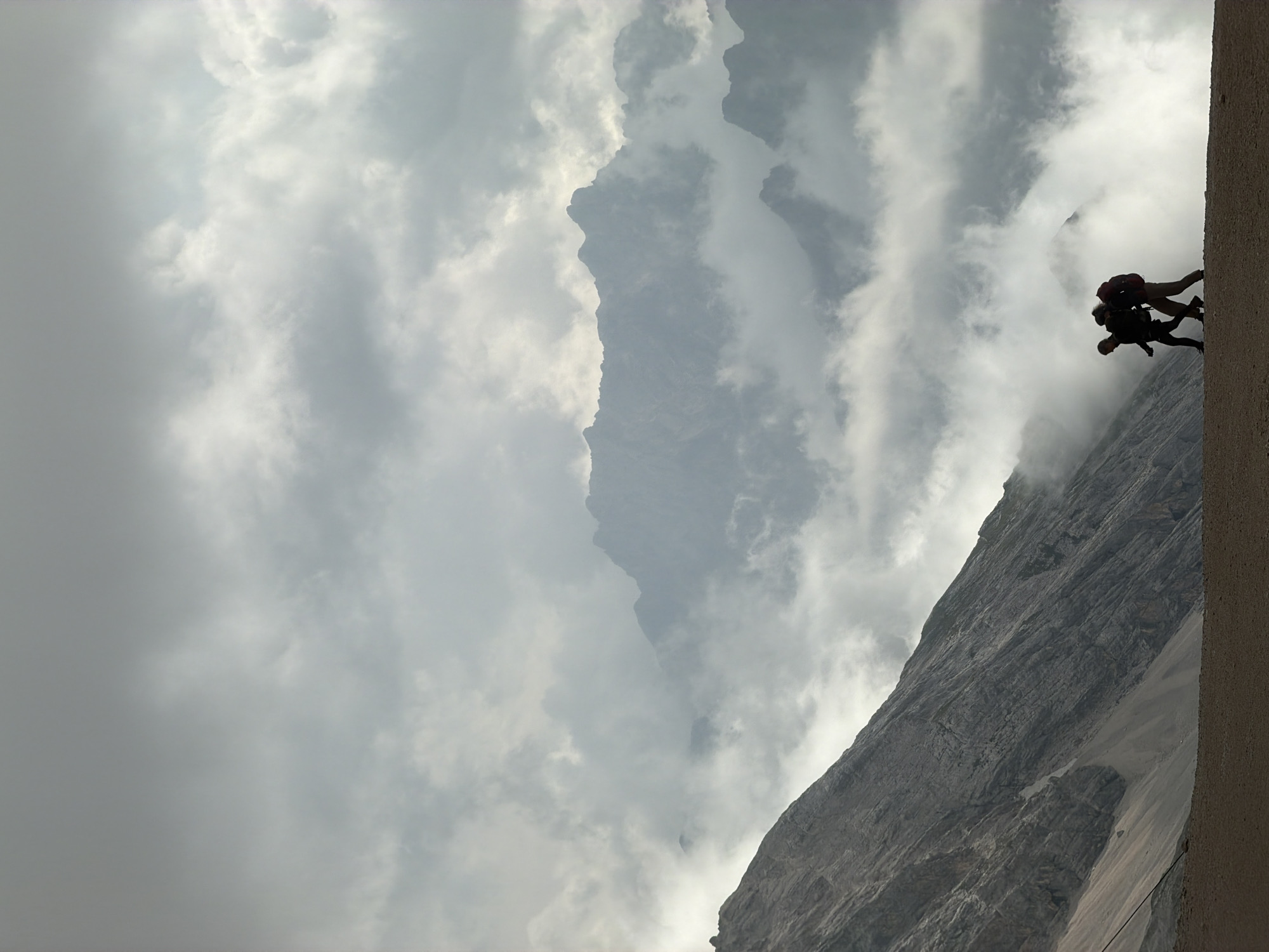 In Wildermieming, Austria, two individuals are captured in silhouette as they walk across a gravelly terrain. The person on the left is wearing dark clothing and has a backpack. The person on the right, also in dark clothing, carries a prominent red backpack. They are walking towards the left of the frame. Behind them, the dramatic landscape unfolds with a steep, rocky mountain slope on the left side of the image. In the background, majestic mountain peaks emerge from a thick layer of swirling clouds, creating a dramatic and atmospheric scene. The sky is overcast with a dense cloud cover.