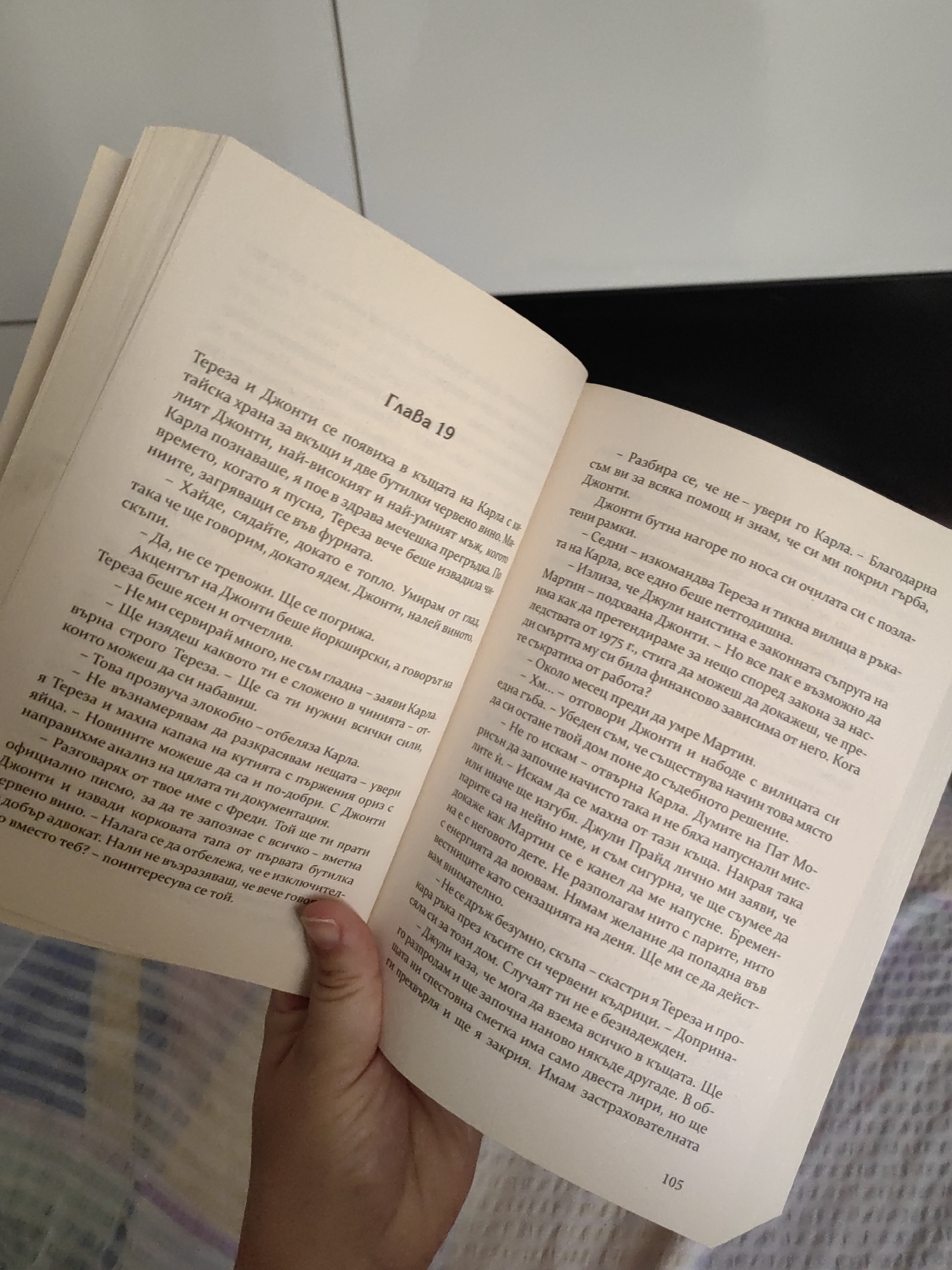 The image shows an open book with text in Bulgarian. The book is a novel, as indicated by the chapter heading "Глава 19" (Chapter 19). The story appears to be about characters named Тереза (Teresa) and Джонти (Djonti), and their interactions with others named Карла (Karla) and Фреди (Fredi). The narrative touches upon themes of money, property, and legal matters, with mentions of inheritance and a lawyer. The book is resting on a dark surface, possibly a table. The background is blurred, but a light-colored wall or ceiling is visible. The image does not contain any information or visual cues that would indicate a specific location in the city of Djerovo, Bulgaria.