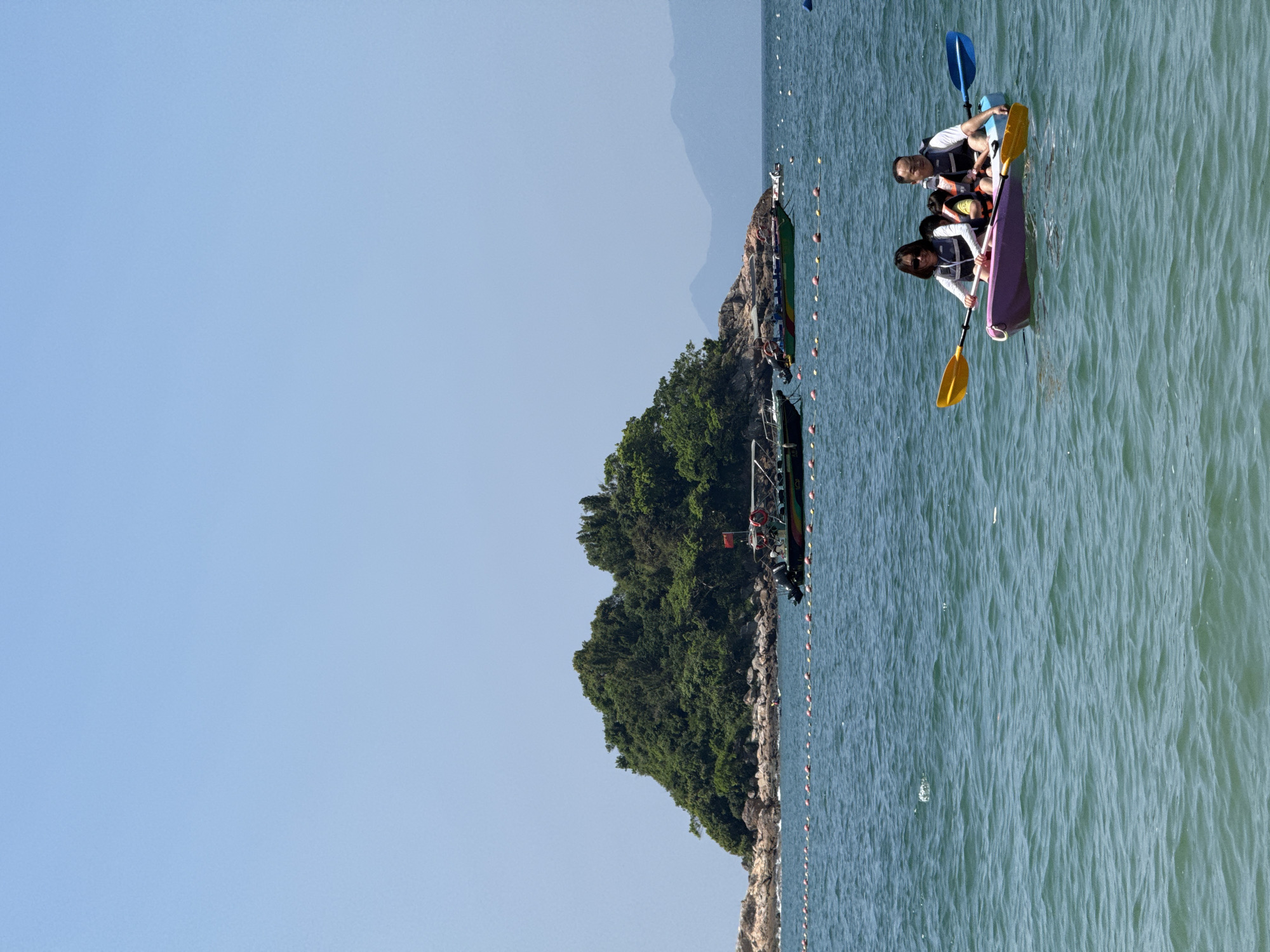 In the foreground, a family of four is kayaking on a calm, blue-green body of water under a clear sky. Two adults and two children, all wearing life vests, are seated in a purple kayak. The adults are paddling with yellow and blue oars. In the mid-ground, a line of red and white buoys stretches across the water. Beyond the buoys, a small, densely forested island rises from the sea, with rocky shores. Several boats are anchored near the island, including a larger tour boat with a red flag. In the background, hazy blue mountains are visible on the horizon. This scene is likely located in an coastal area within Longgang District, China, known for its natural beauty and recreational water activities.