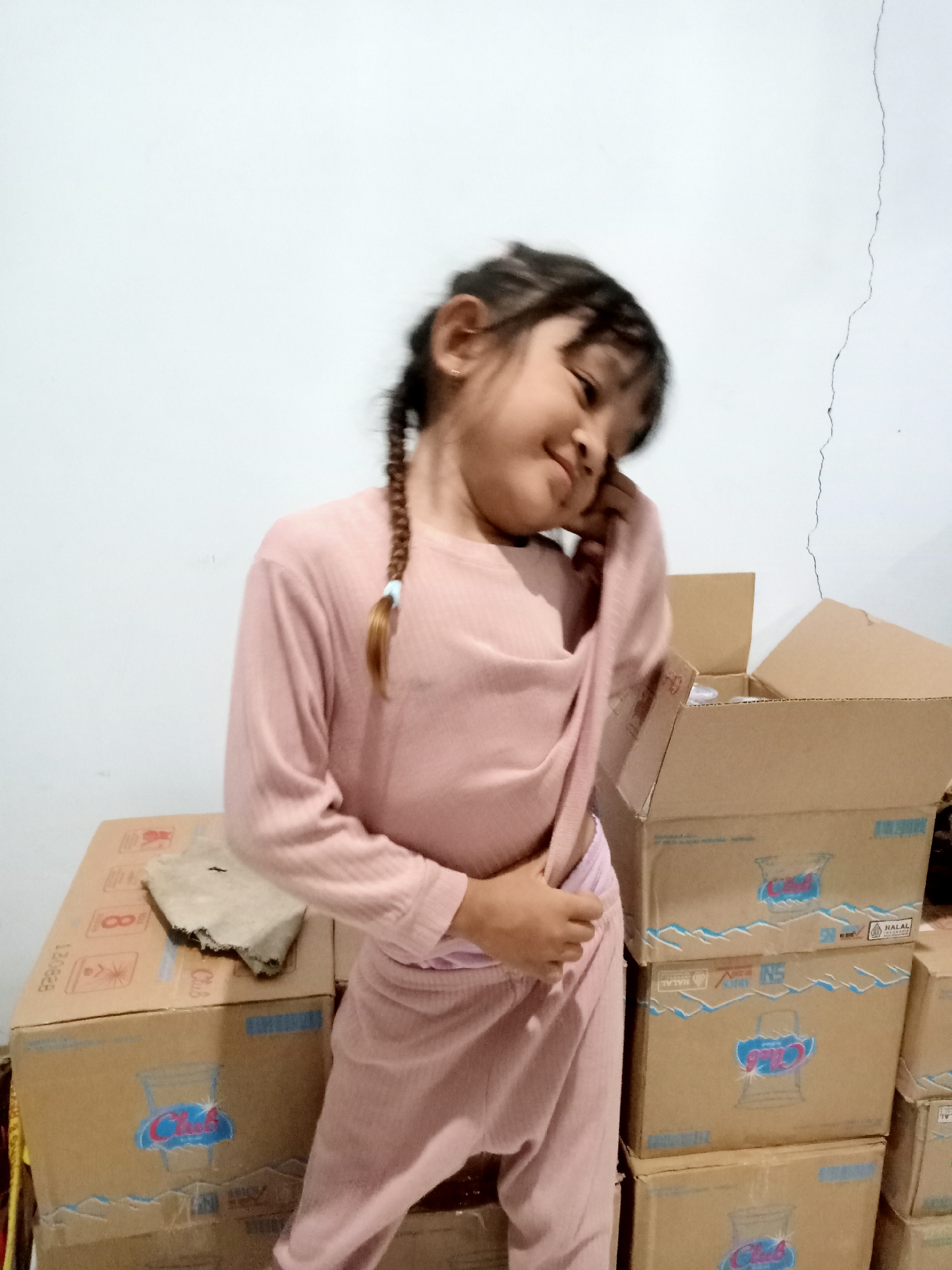 A young girl with braided hair, wearing a pink long-sleeved shirt and pink pants, stands in front of several cardboard boxes. She is looking down and to her right, with her right hand touching her face. The background is a plain white wall with a vertical crack. The boxes appear to be stacked and some have branding with blue lettering and images of cups. The scene suggests an indoor environment, possibly a storage area or a home in Simo, Indonesia.