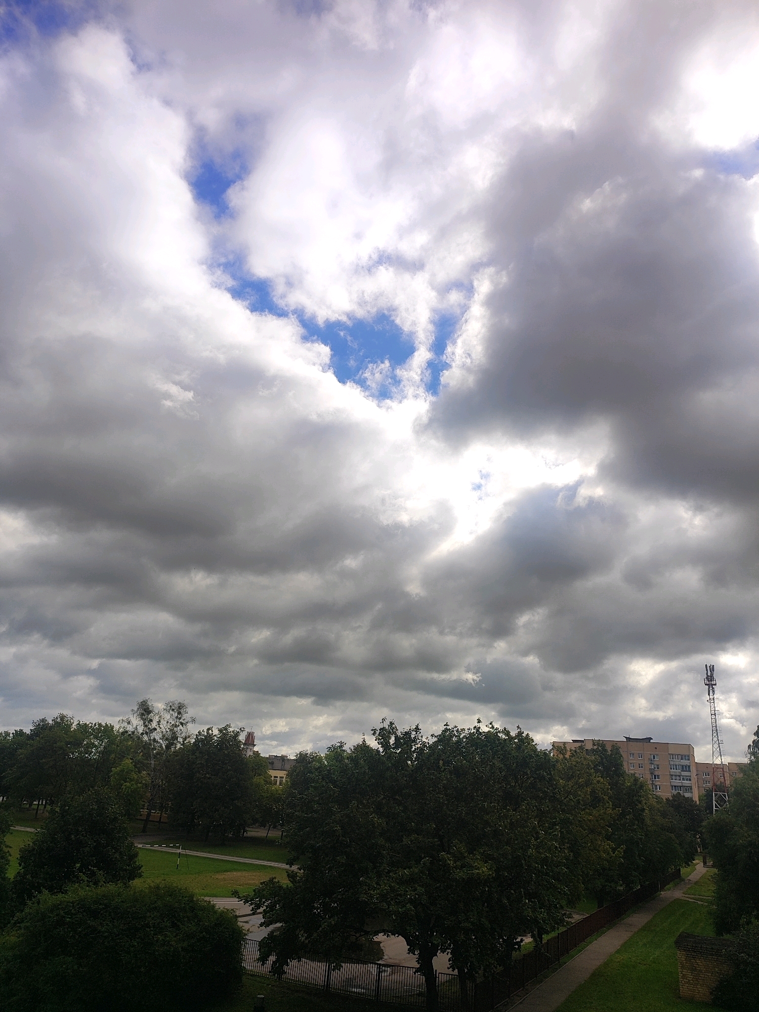 An aerial view of a city park with lush green trees and pathways is depicted under a dramatic sky filled with large, dark, and white clouds. In the background, a multi-story apartment building with visible windows stands, and a tall cell tower is prominent against the clouds. The scene is devoid of people. The setting appears to be a residential area with a park, likely Hrodna, Belarus, given the architectural style and general landscape.