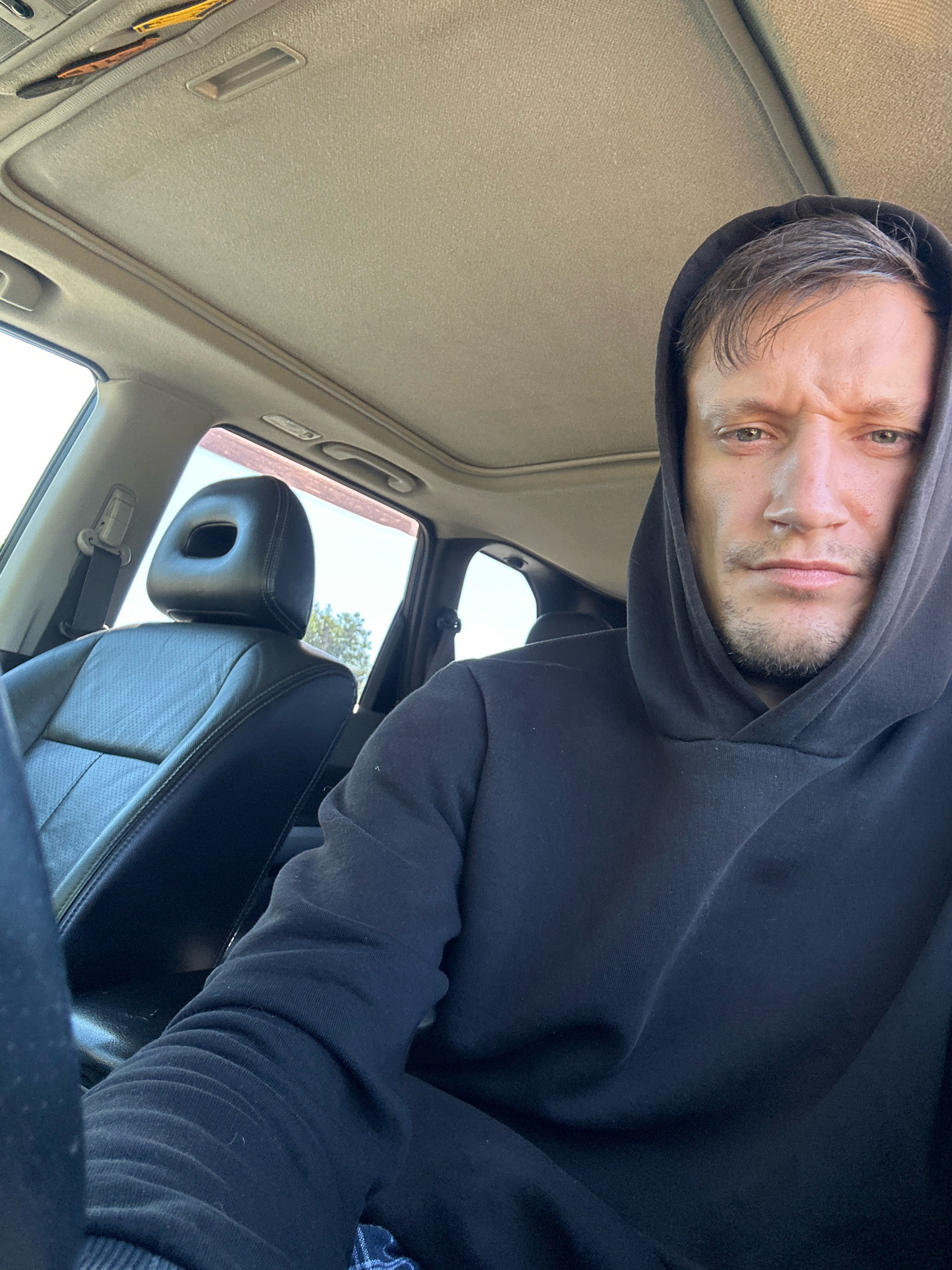 A man in a black hoodie is sitting in the driver's seat of a car, looking at the camera. The car has a sunroof open, and the interior is visible, including a black leather seat. The man has short, dark hair and appears to be in his late 20s or early 30s. The image is taken from a slightly high angle, looking down at the man. The environment outside the car is not clearly visible, but sunlight suggests it is daytime. The image does not provide any information about the specific location within the city of Makiv, Ukraine.