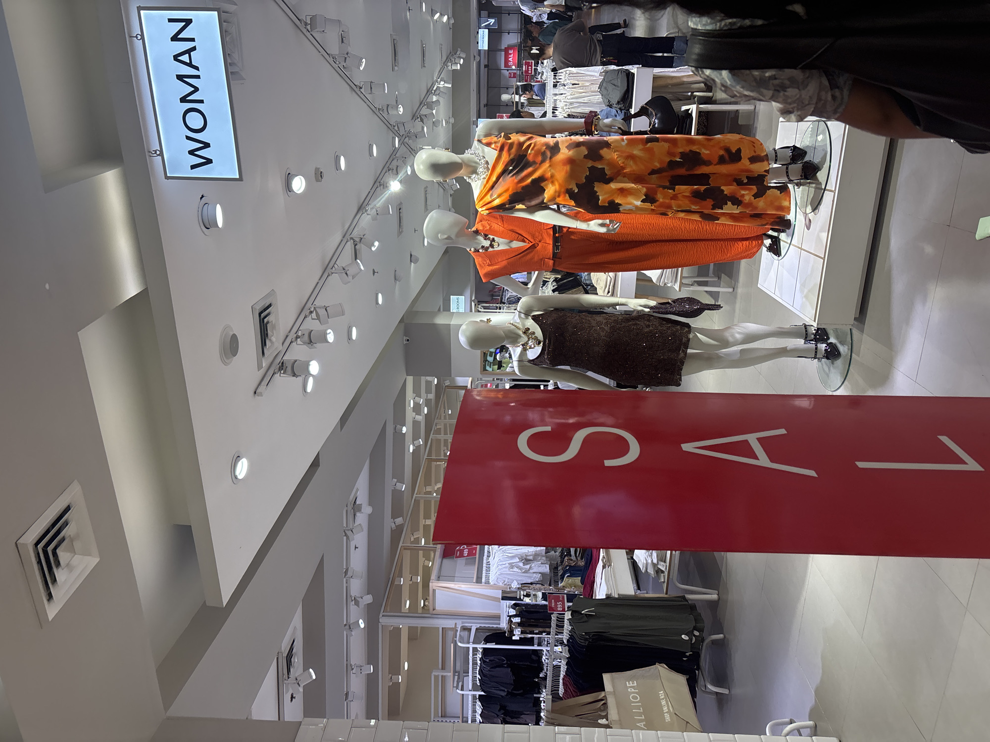 The image shows the interior of a women's clothing store, likely a department store or a chain retailer, with bright overhead lighting and white tiled floors. A large red sign with white lettering, partially obscured but spelling out "S A L" from "SALE", is prominently displayed in the foreground. To the right of the sign are three mannequins dressed in women's fashion. The first mannequin on the left wears a sequined brown dress. The second mannequin is dressed in an orange blazer and matching pants. The third mannequin, on the far right, wears a patterned orange and brown sleeveless dress. Several racks of clothing are visible in the background, along with signage indicating "WOMAN" and "FITTING ROOMS". The store appears to be busy, with other shoppers visible in the background. The presence of "WOMAN" signage and the styles of clothing suggest this is a women's fashion section. This scene is consistent with a retail environment found in a mall in Davao City, Philippines, such as those located in Abreeza Mall or SM Lanang Premier, where various clothing brands and department stores are situated.