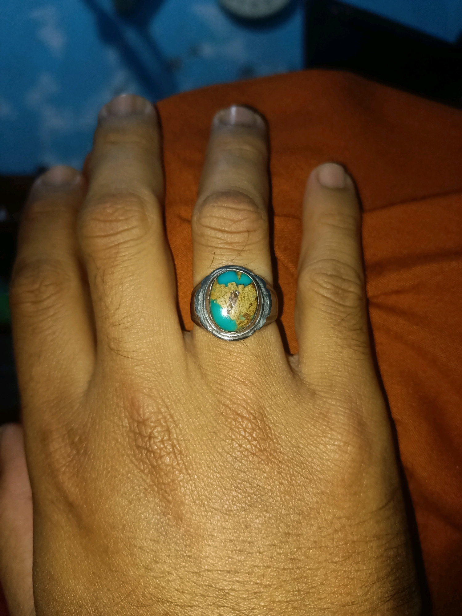 An extreme close-up shot shows a person's hand wearing a ring with a turquoise-colored stone. The hand is resting on an orange surface. The background is blurred and appears to be a blue wall with some white patterns. The image is lit from above and to the left, casting shadows on the hand. The image location is stated as Wadeng, Indonesia.