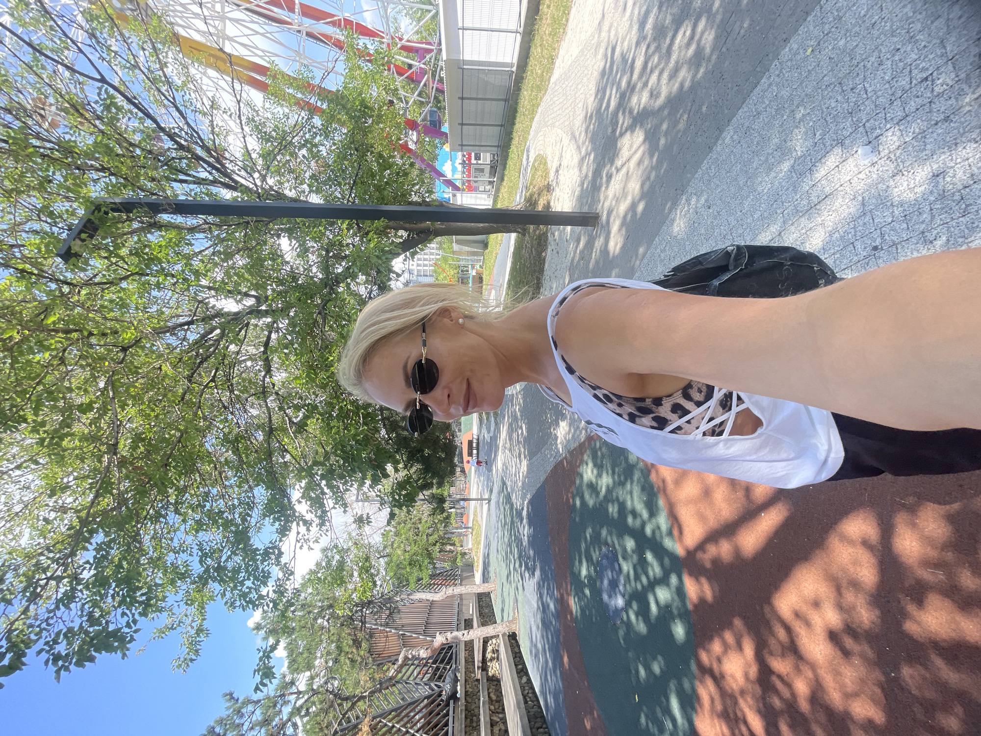 A woman with blonde hair and sunglasses takes a selfie in an outdoor amusement park. She is wearing a white tank top with a leopard print bra visible underneath and a black backpack. The background shows a ferris wheel, trees, and a paved walkway. The image appears to be taken on a sunny day in Novorossiysk, Russia.