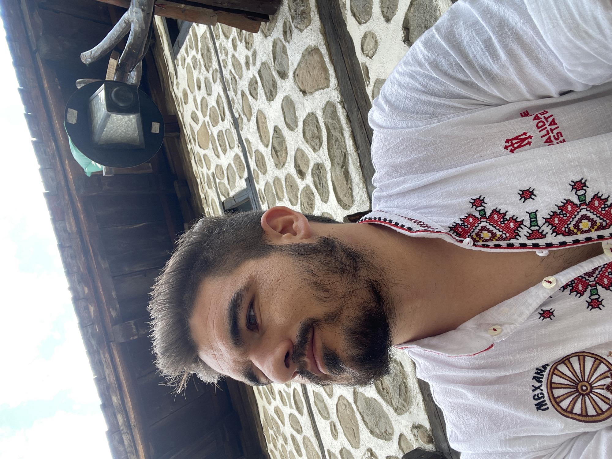 A low-angle, eye-level shot captures a man from the chest up, positioned slightly to the left of the frame, against a rustic stone and wood building in an unknown location. The man has a dark beard and mustache, dark eyes, and dark hair styled upwards. He is looking off to his left (viewer's right). He wears a long-sleeved white shirt with a textured fabric.
The shirt features extensive embroidery:
* On the left chest, the Cyrillic word "МЕХАНА" (Mekhana) is embroidered in dark brown above a stylized wagon wheel design in brown and gold.
* The front placket and collar are adorned with red, black, and yellow geometric and floral patterns.
* On the right chest, a red emblem with interlocking "M" shapes is visible above the text "ALIDAE ESTATE" (the initial 'V' is partially obscured, suggesting "VALIDAE ESTATE") in red.
* The collar and placket edges have a thin red trim, and the shirt is fastened with white, two-hole buttons.
Behind the man, a building constructed with irregular light-colored stones set in thick white mortar is visible. Above the stone wall, dark wooden beams indicate a roofline or overhang. In the upper right background, an old-fashioned, dark-colored outdoor lantern with a textured glass face is mounted on a gnarled wooden support beam. The sky above is light blue with scattered white clouds, suggesting daylight conditions.