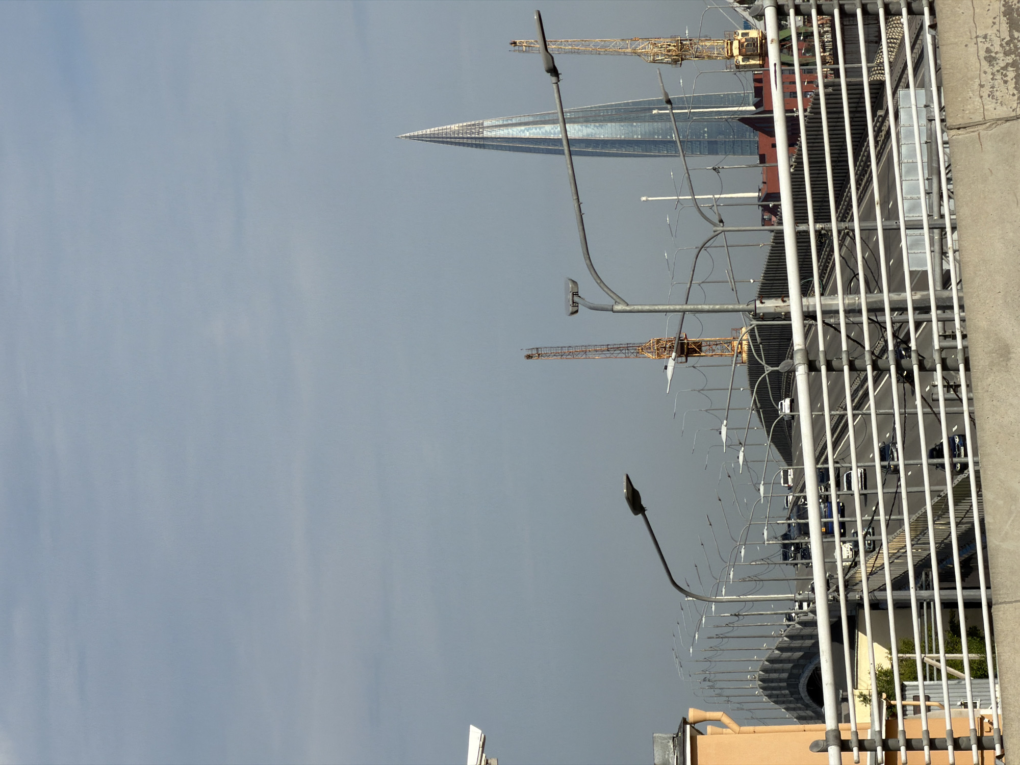 The image shows a city skyline in Saint Petersburg, Russia, dominated by the Lakhta Center skyscraper, a tall, modern building with a pointed roof. To the left of the skyscraper are two construction cranes, suggesting ongoing development in the area. In the foreground, a multi-lane highway bridge spans across a body of water, with cars visible driving on it. Streetlights line the bridge, and a security camera is mounted on a pole on the left side of the image. The sky is overcast, with a light blue hue. The overall scene depicts a busy urban environment with both existing infrastructure and ongoing construction.