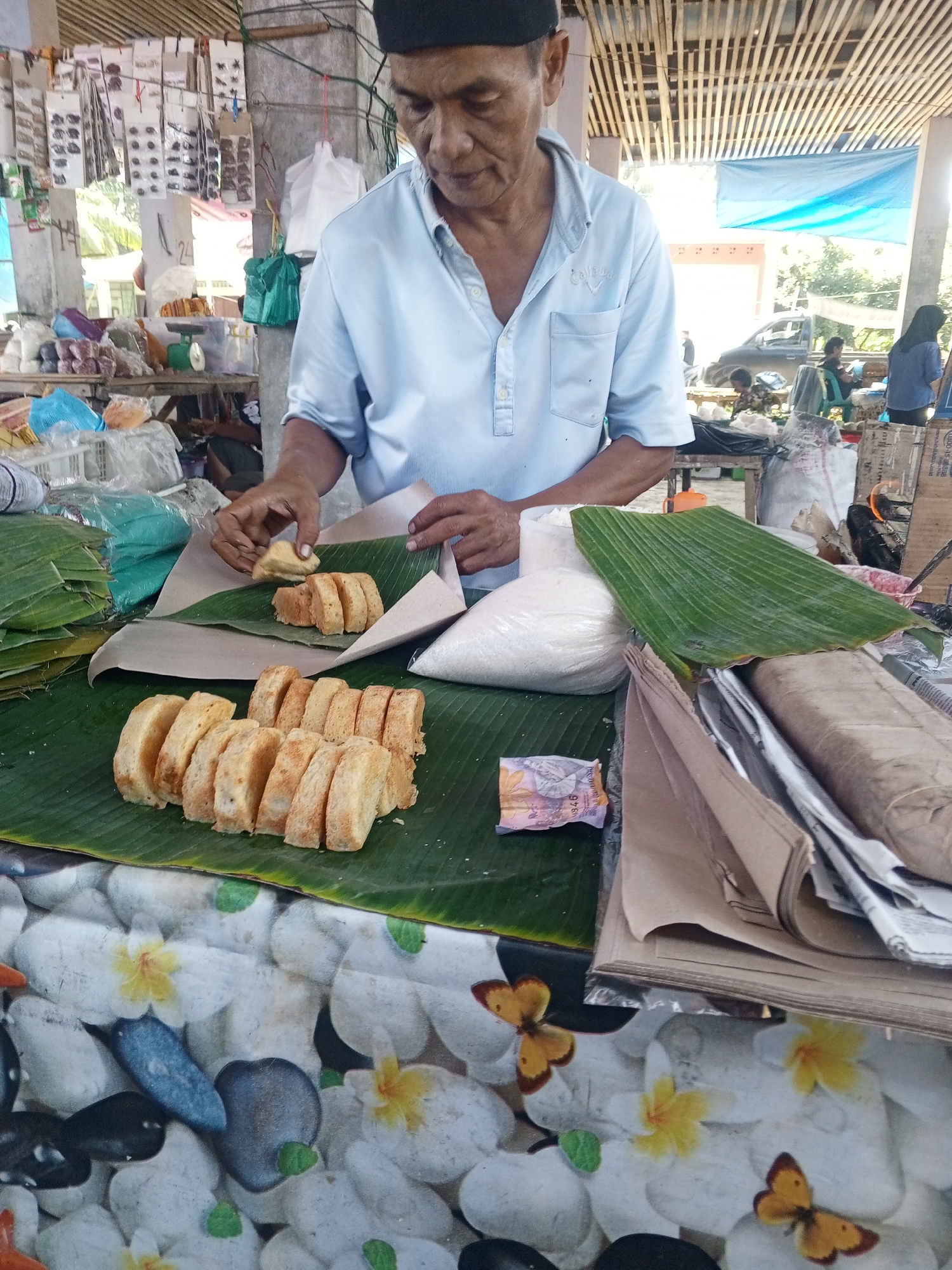 An older Indonesian man with dark hair and a light blue collared shirt is standing behind a market stall in Talun Kenas, Indonesia. He is wearing a black cap. The man is in the process of arranging small, sliced pieces of a light brown, sweet-looking baked good on a large green banana leaf. Another banana leaf serves as a barrier behind the baked goods. In front of him is a white, granular substance in a clear plastic bag, and a folded piece of brown paper. To the left of the baked goods, there are more green banana leaves stacked up. The stall is set up outdoors, with a bamboo-like structure overhead, and a blue tarp visible in the background. Other market stalls and some vehicles are also present in the background, suggesting a bustling market environment. The table the man is working at is covered with a white cloth featuring a pattern of white pebbles and yellow and orange flowers, with a few monarch butterflies also depicted.