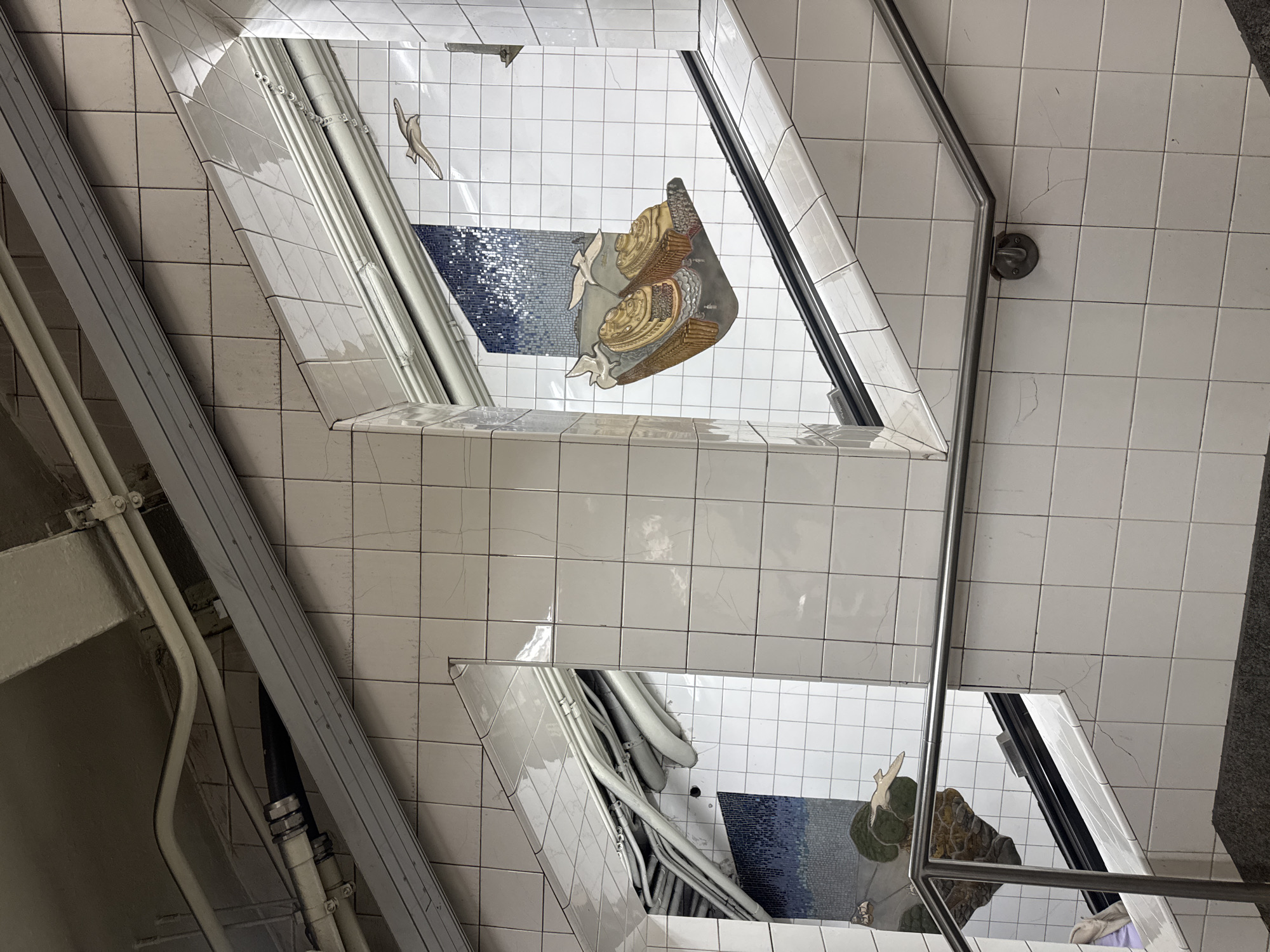 This image captures an angled view within the Grand Central - 42nd Street subway station in New York City, United States of America. The scene depicts a stairwell or ramp, predominantly surfaced with square, glossy white ceramic tiles that exhibit visible grout lines and some hairline cracks.
Exposed utility pipes and conduits, primarily light grey and off-white, run diagonally across the upper sections of the image, with a dark pipe visible in the upper left. A large, grey, rectangular metal housing or fixture runs parallel to the slope of the wall in the upper right.
The central focus is a multi-panel mosaic artwork, "Oyster Pail" by Laura Battle. On the right, a vibrant mosaic depicts two large, golden-brown oyster shell barges filled with smaller shells, positioned against a backdrop of dark and light blue mosaic tiles representing water or sky. Two white seagulls are shown in flight above these elements. A partial reflection of similar mosaic art, including a green-brown landform and blue background, is visible on a reflective surface or column on the left side.
A polished metal handrail descends diagonally on the right, attached to the tiled wall by a circular bracket. A segment of a similar handrail is present on the lower left. Dark grey, granular steps are visible at the bottom of the frame. The area is illuminated, causing reflections on the glossy tile surfaces. No individuals are present in the immediate view.