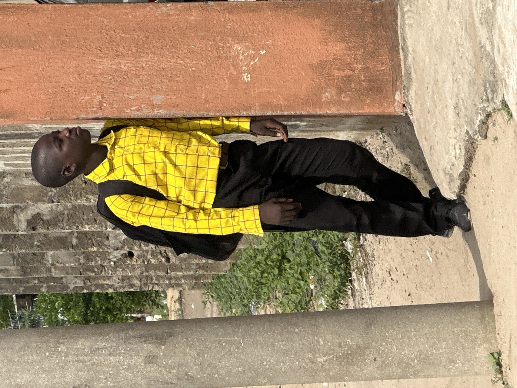 This full-body outdoor shot, taken in Badagry, Nigeria, features a young person, likely male, standing under bright daylight. The subject has dark skin and a short, dark haircut. He is dressed in a long-sleeved, collared yellow shirt with a distinct black grid pattern, black trousers, and black shoes, carrying a black backpack on his shoulders. His head is tilted slightly upwards, and he appears to be looking towards his upper right with a neutral to contemplative expression.
The setting consists of various architectural elements. The person stands on a light-colored concrete or paved surface that shows some minor wear and scattered green foliage. To the right of the frame, a tall, reddish-brown textured wall or facade dominates, possibly made of aged concrete or brick. On the left, a grey concrete pillar or vertical structure is visible. Behind the subject, a rough-textured grey wall or barrier is partially covered by dense green leafy vegetation. The bright, even illumination suggests the photograph was taken during the day, likely mid-morning or afternoon, in sunny weather. There is no visible text in the image. The scene conveys a typical urban or semi-urban environment in a tropical region.