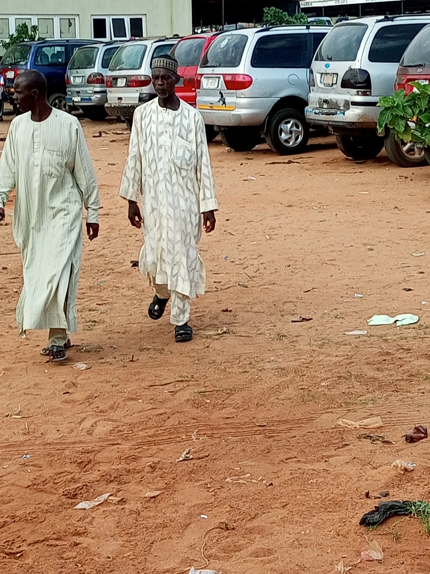 Two men are walking on a dirt path. The man on the left is wearing a light green traditional robe and sandals. The man on the right is wearing a white traditional robe with a patterned design and black sandals. They are walking away from the camera. In the background, several vehicles are parked, including what appears to be a silver minivan, a red minivan, and a blue minivan. The area looks like a parking lot or an open space next to a building. The ground is covered in reddish-brown dirt with some scattered debris, suggesting a dry, possibly dusty environment. The lighting suggests it is daytime. The scene appears to be outdoors in a somewhat rural or less developed area, possibly in Gombe, Nigeria, given the context. There is no visible text.