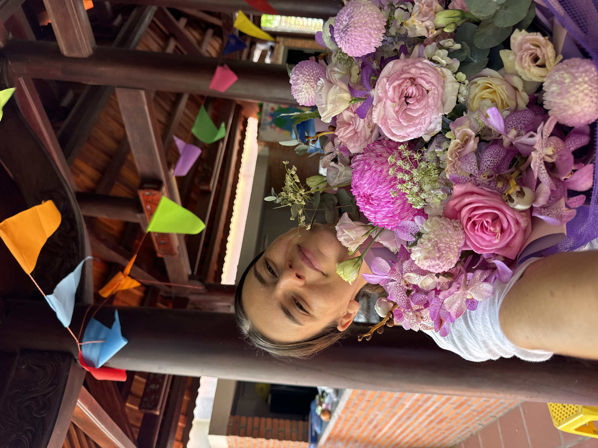A young woman with dark hair pulled back is holding a large bouquet of pink and purple flowers, including orchids and roses, directly in front of her face, obscuring much of her body. She is wearing a white t-shirt. The background suggests an outdoor covered structure with wooden beams and columns. Colorful triangular flags are strung across the ceiling. To the left, a brick wall and a yellow chair are visible. The overall impression is a celebratory or gift-giving scene, possibly in a park or cultural site in Mũi Né, Vietnam.