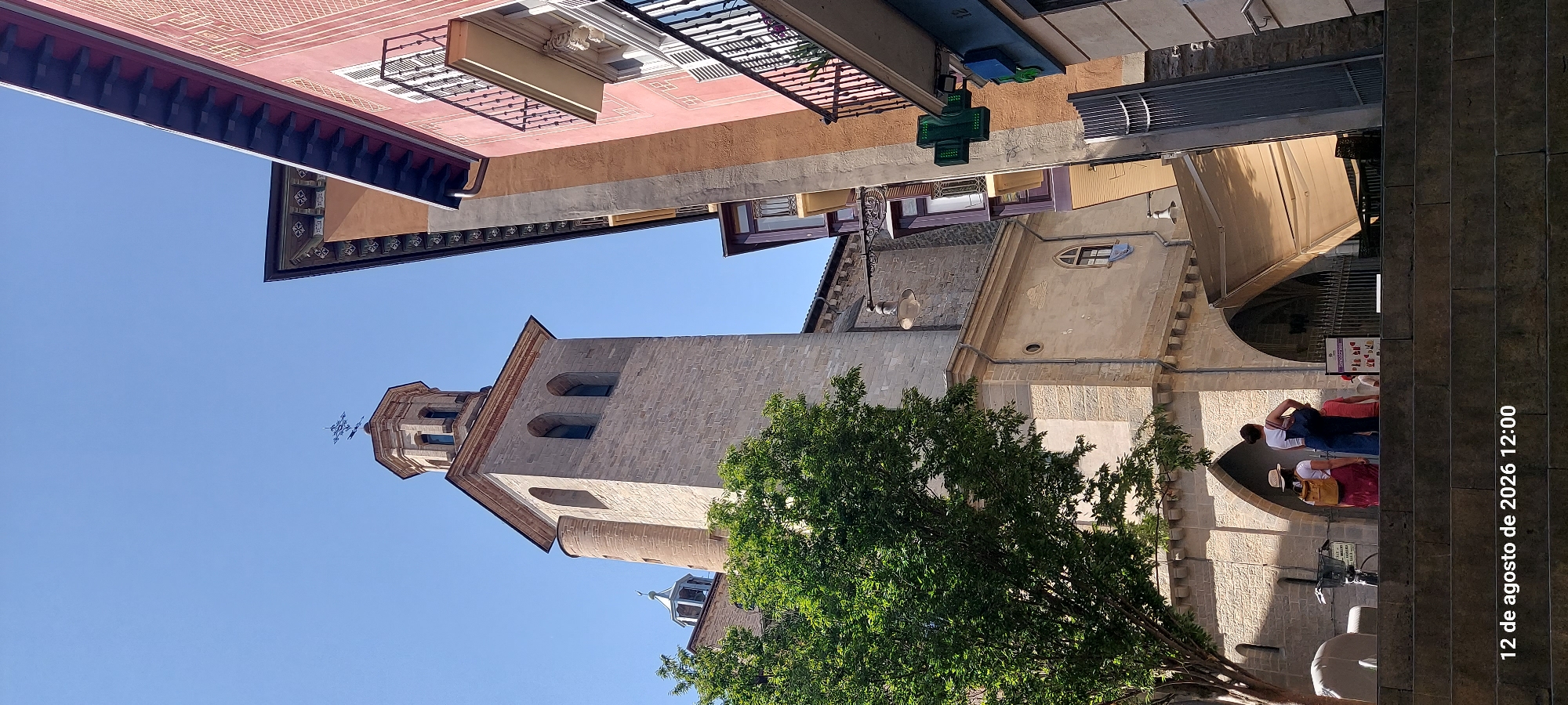 This image captures a street scene in Pamplona, Spain, featuring a prominent stone church tower and adjacent buildings under a clear blue sky. The church tower, made of light-colored stone blocks, has a square base that transitions to an octagonal upper section topped with a decorative cross. A round tower is attached to the left side of the main tower. In the foreground, two people are standing on a stone staircase, facing away from the camera. One person wears a straw hat and a yellow backpack, while the other has dark hair and a blue bag. To the right of the church, buildings with balconies and colorful facades are visible. A bright green pharmacy cross is mounted on one of the buildings. The overall environment appears to be an urban setting with historical architecture. The date and time stamp "12 de agosto de 2026 12:00" is visible at the bottom left of the image.
