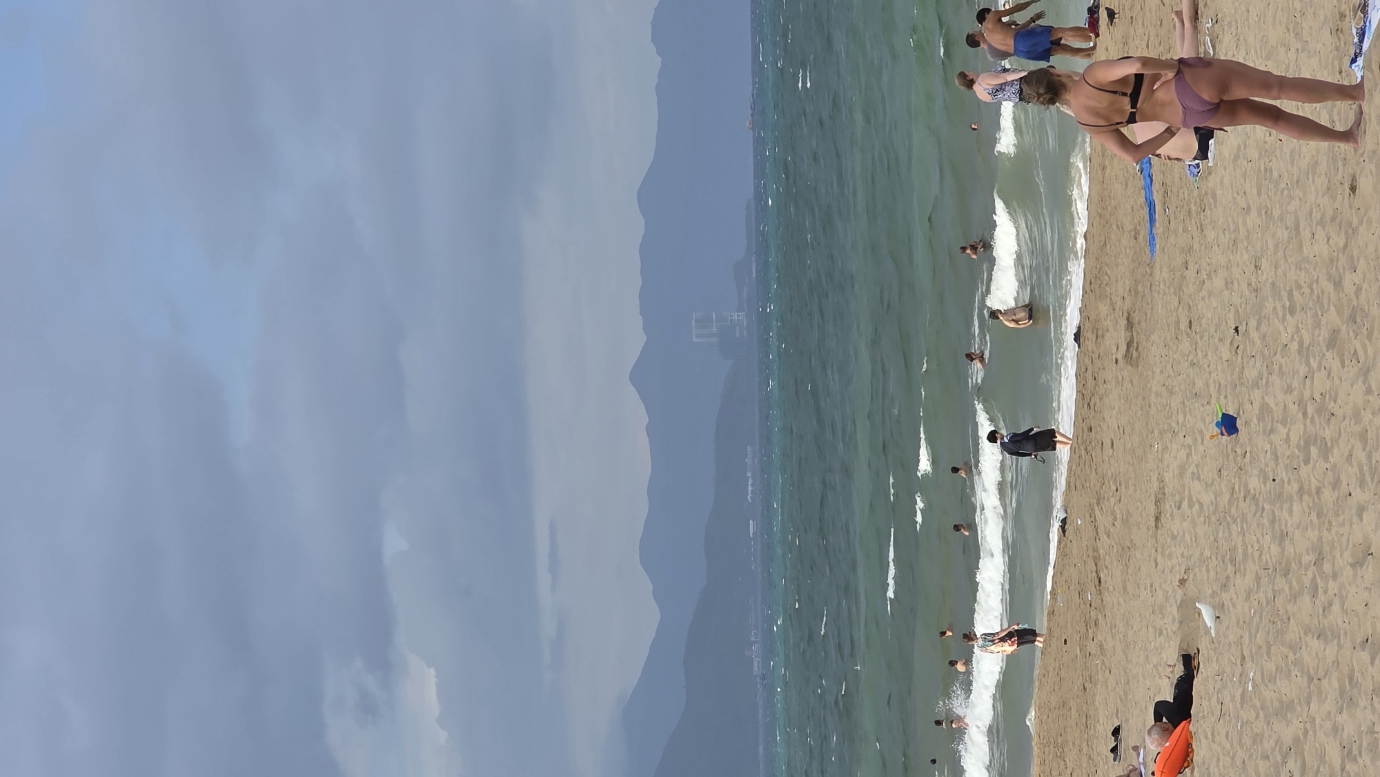 The image depicts a crowded beach in Nha Trang Ward, Vietnam. The sky is overcast, and mountains are visible in the background. The sea is choppy with small waves. Numerous people are on the beach, some swimming or wading in the water and others lounging on the sand. A woman in a bikini stands on the sand near the water's edge. A man in dark clothing stands in the shallow water. Other individuals are scattered along the shoreline. The sand appears light brown. There is an orange object and a blue pail visible on the beach.