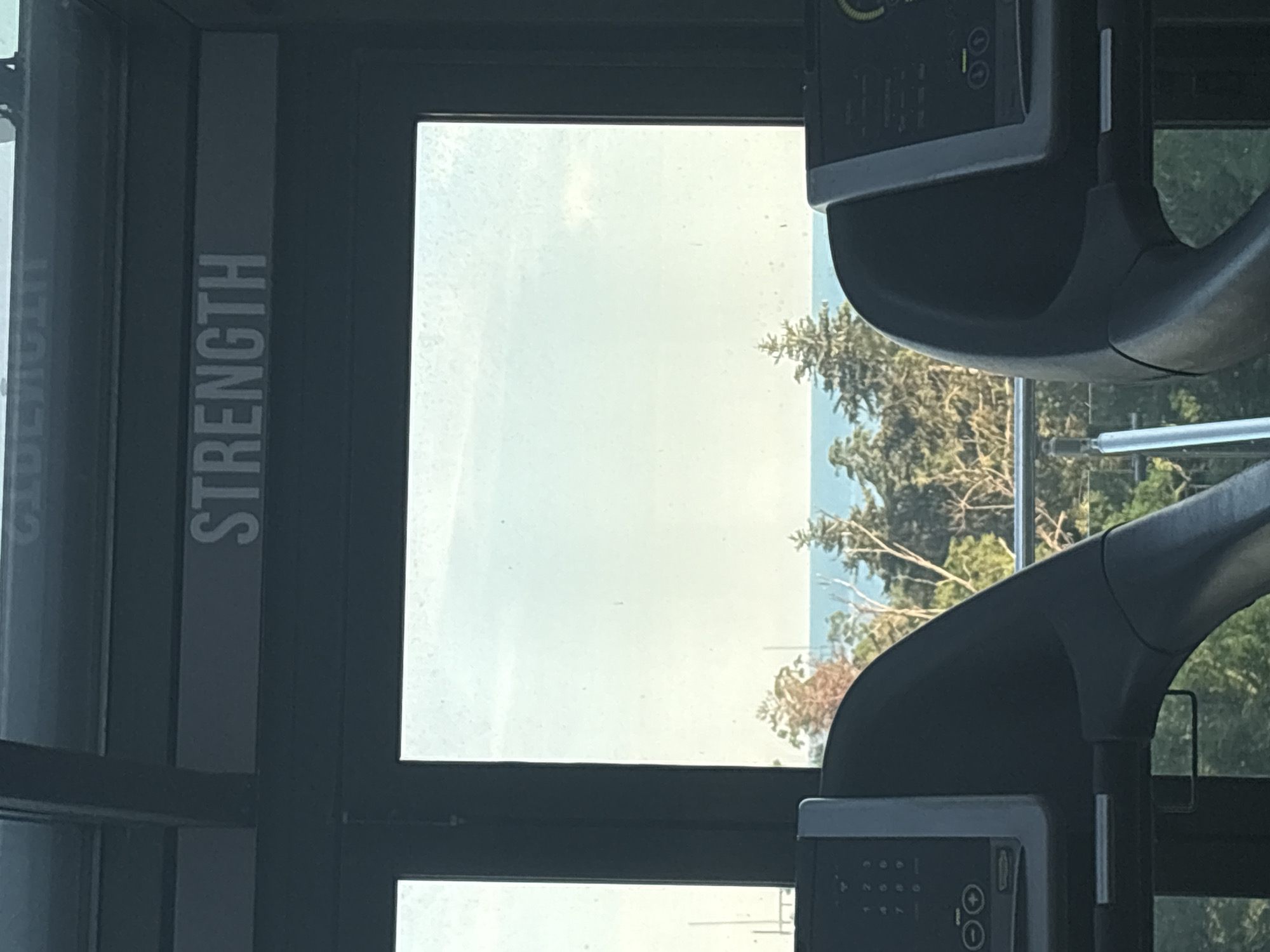 The image shows a view through a window of a gym, looking out at a body of water and trees. In the foreground, the top consoles of two treadmills are visible, one on the left and one on the right. The console on the left has a numerical keypad and speed control buttons. Above the window, a sign reads "STRENGTH" in white capital letters on a gray background. The window reveals a clear sky with a hint of clouds, and a blue body of water beyond a line of green trees. The scene appears to be a gym with a scenic view.