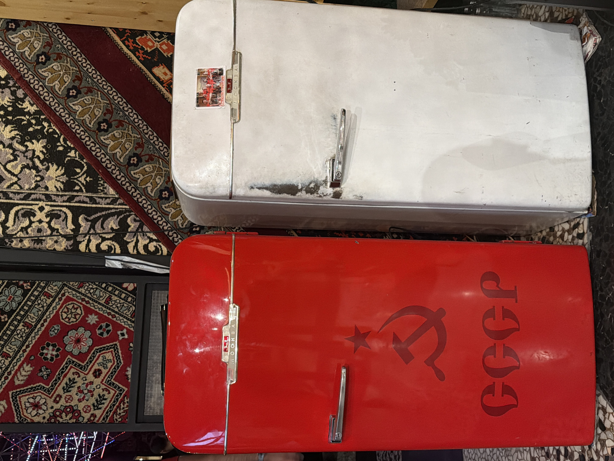 The scene depicts two upright, vintage-style refrigerators positioned side-by-side in an indoor setting in Sofia, Bulgaria. The refrigerator on the left is bright red with a glossy finish. Its upper section features a chrome emblem with the Cyrillic inscription "MOCKBA" (MOSCOW). A chrome horizontal handle is affixed to the left side of its door. On the lower half, a darker red stencil depicts a hammer and sickle symbol surmounted by a star, with the Cyrillic letters "CCCP" (USSR) below it. This refrigerator appears clean and reflective.
The refrigerator on the right is off-white or light grey and exhibits signs of wear and discoloration, including a prominent dark vertical streak on its left side near the handle. It also features a chrome emblem with the "MOCKBA" inscription. A small, rectangular magnet or sticker is affixed above this emblem, displaying a group of approximately five to six individuals in red attire. A chrome horizontal handle is present on the left side of its door.
The background consists of decorative rugs or tapestries hanging on the wall. To the upper left, a rug with red, black, and beige geometric patterns is visible. To the upper right, another rug with darker, possibly black and gold, floral or scroll patterns is present. Below the left rug, a dark rectangular object, possibly a speaker or vent, is partially visible. Further to the left, blurred multi-colored lights suggest an illuminated backdrop. The floor is composed of dark and light-toned mosaic-patterned tiles. A blurred human silhouette is faintly discernible on the far left, behind the red refrigerator.