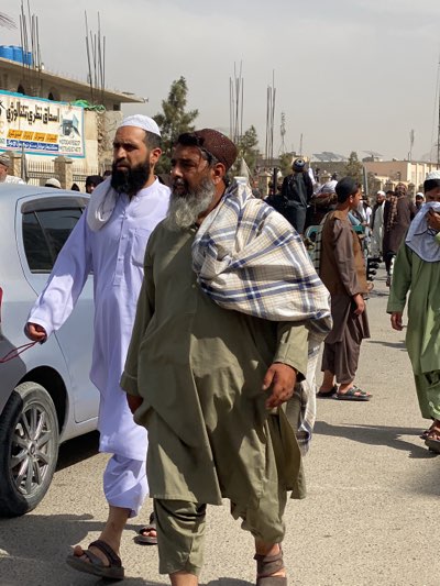 The image shows a group of men walking in Kandahar, Afghanistan. Two men are in the foreground, one wearing a white thobe and skullcap, the other a brown hat and a plaid shawl over a green outfit. They are walking on a paved road alongside a silver car. More men are visible in the background, some wearing traditional clothing. Buildings line the street, and utility poles are visible. The sky is hazy, and the scene appears to be outdoors during daylight.