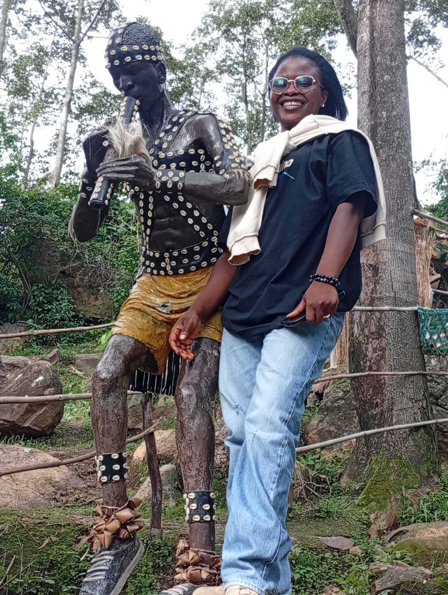 A woman wearing glasses, a black t-shirt, and light blue jeans poses next to a life-sized dark statue of a man playing a flute. The statue is adorned with shells and wears a yellow skirt and black and white striped fabric. The background features trees, rocks, and lush greenery. The woman has a light-colored sweater draped over her shoulders and is holding a phone in her right hand.