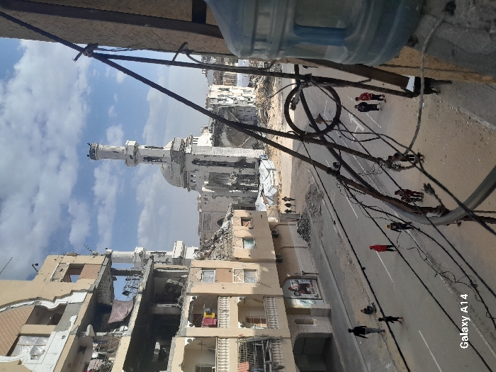 This image captures a wide view of a heavily damaged urban environment, specifically depicting the area around the Al-Fara Mosque in Khan Yunis, Palestinian Territory, under a partly cloudy sky.
In the background, the prominent white Al-Fara Mosque stands with its large dome and tall, multi-tiered minaret; both structures exhibit visible signs of damage. Surrounding the mosque are extensive areas of rubble and skeletal remains of buildings. Several temporary shelters constructed from white tarpaulin are visible amidst the debris further in the background.
On the left side of the midground, a multi-story building shows significant structural damage, with large sections of its outer walls collapsed, exposing interior rooms where laundry is visible hanging on lines.
A paved asphalt road traverses the midground. Approximately ten individuals, predominantly children, are present on the street. On the left side, a man in dark attire walks away from the viewer, while two children sit on the roadside curb. Another child walks further down the street. In the center, a child in a red top stands alone. On the right side, a group of five children and one adult or older teenager are gathered near the curb. The street surface is dusty and shows signs of debris.
In the immediate right foreground, a partial view of a large, blue plastic water dispenser bottle is visible. Numerous tangled black cables or wires and several metal support rods crisscross the right side and mid-foreground, partially obstructing the view of the street below.