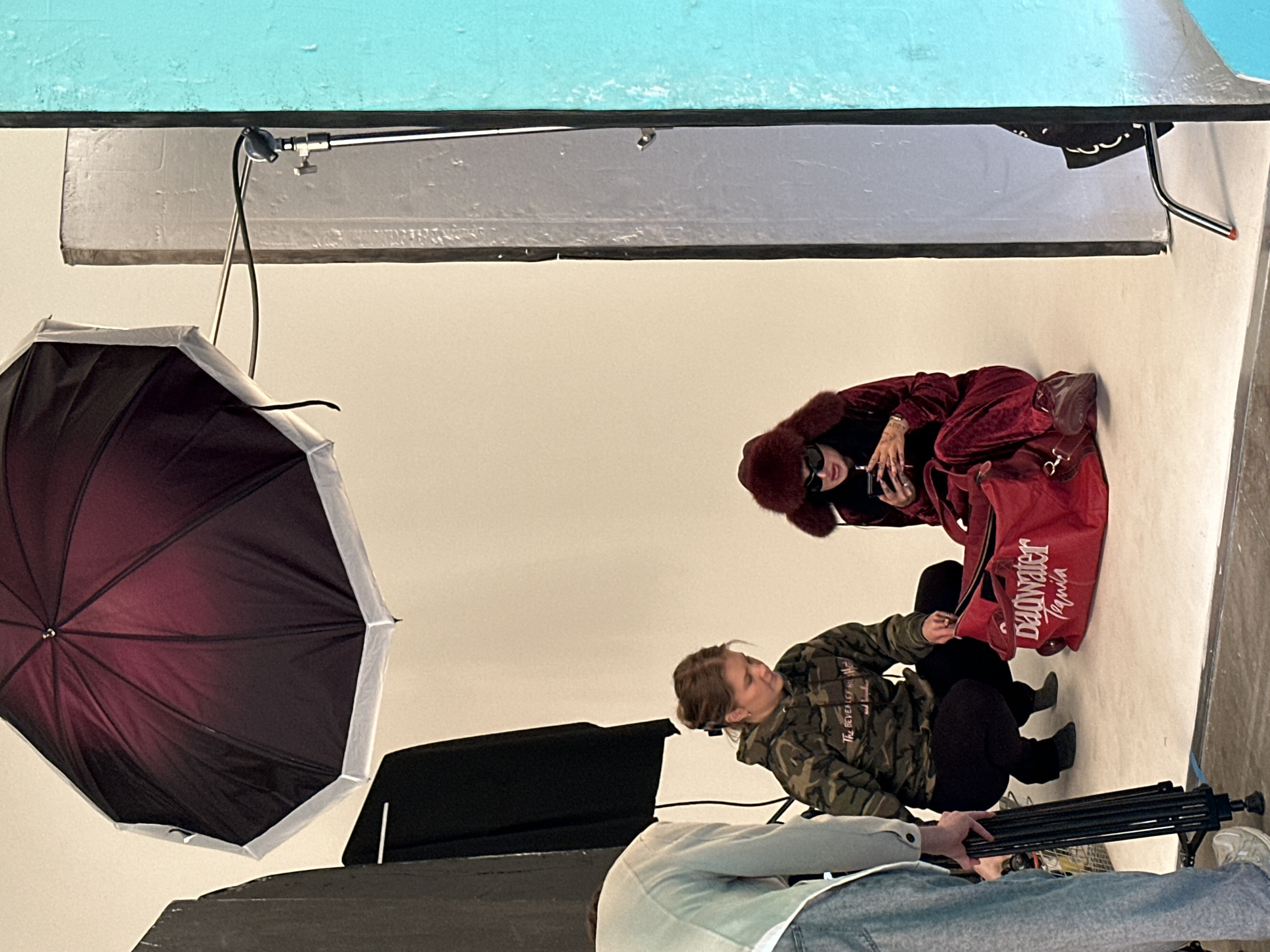 A behind-the-scenes image captures a photoshoot setup. A woman in a fur-trimmed red suit and sunglasses poses with a large red bag labeled "Dadwater Tequila." Another woman in a camouflage jacket crouches nearby, adjusting equipment. An open black and red umbrella serves as a light modifier, positioned to the left. Studio lighting and support structures, including a tripod, are visible, suggesting a professional studio environment.