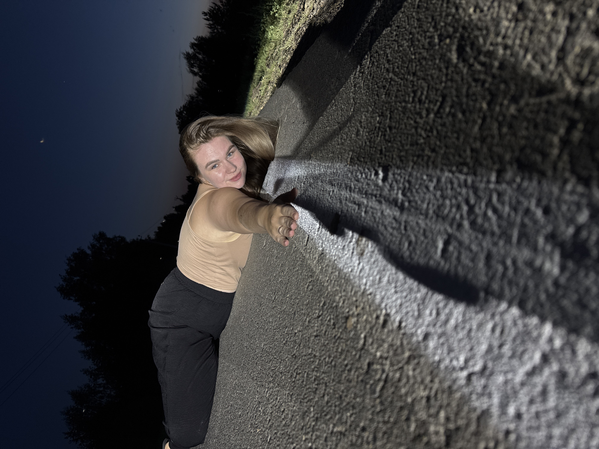 A young woman with blonde hair is posing on a concrete structure at night. She is wearing a tan tank top and black sweatpants. She is holding the edge of the structure with her right hand. The background shows trees and a dark sky with a crescent moon. The location is presumably in or near Ольховка, Russia.