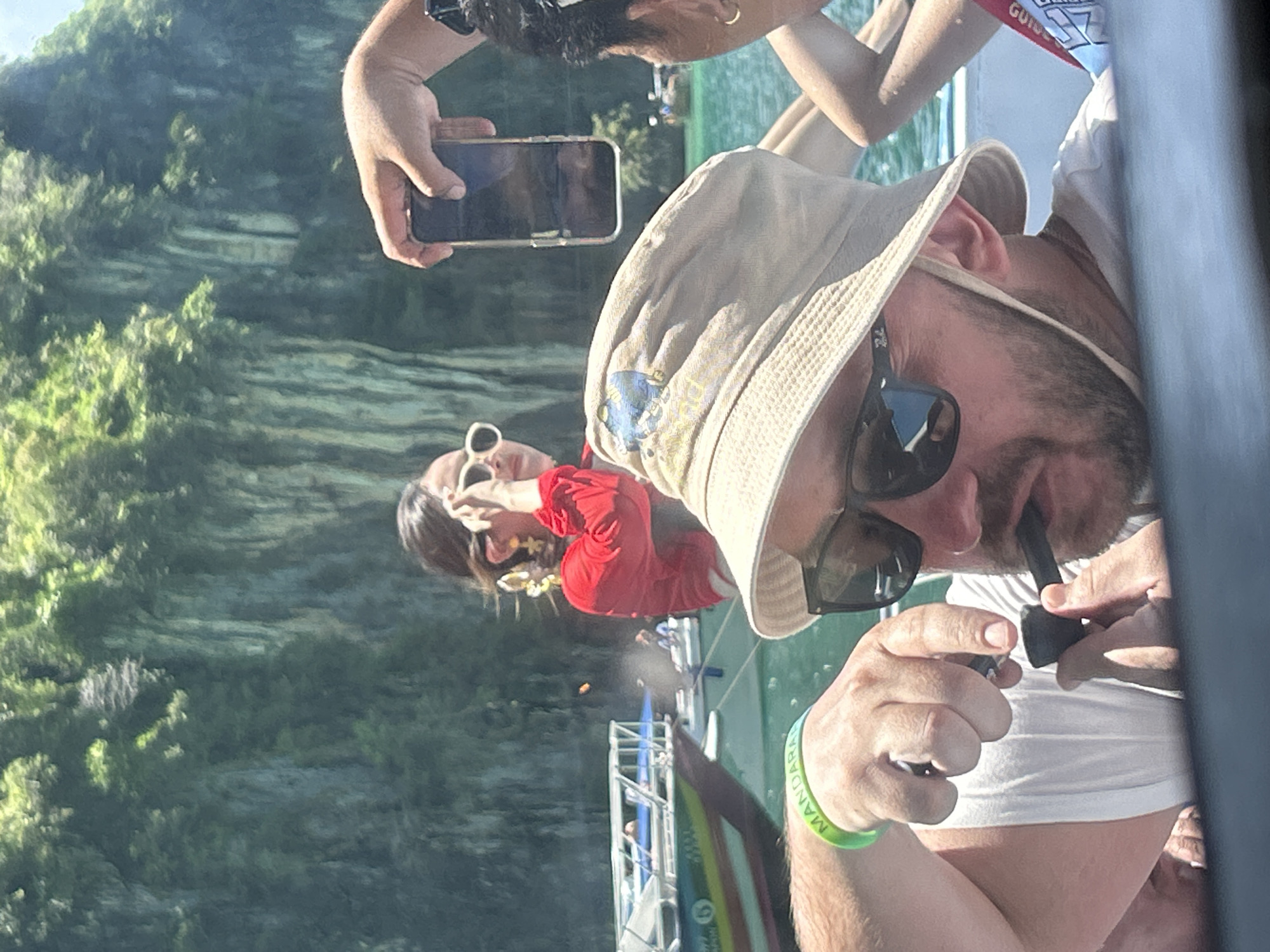 A high-angle daylight photograph captures a group of individuals on a boat against a backdrop of towering limestone karsts covered in dense tropical foliage, characteristic of the marine environment accessible from Ao Nang, Thailand. In the foreground, a light-skinned male with a beard and dark sunglasses, wearing a light-colored bucket hat with a chin strap, holds a small black pipe to his mouth with his right hand. A neon green wristband displaying "MANDARA" in white text is visible on his left wrist. To his right, a partially visible individual with dark hair and a gold hoop earring holds a foldable smartphone vertically, pointed towards the background, seemingly capturing the scene. In the midground, to the left, a female with dark hair wears a bright red long-sleeved top and large, intricate gold earrings. She wears white-framed sunglasses, which she appears to be adjusting with her right hand, while looking forward. The boat is on turquoise-green water. In the background to the left, another longtail-style boat, predominantly white and blue with colored stripes and the number "6" visible on its hull, is anchored or moving slowly. Further in the distance, additional boats are visible on the water, with the green cliffs rising prominently behind them under a bright sky.