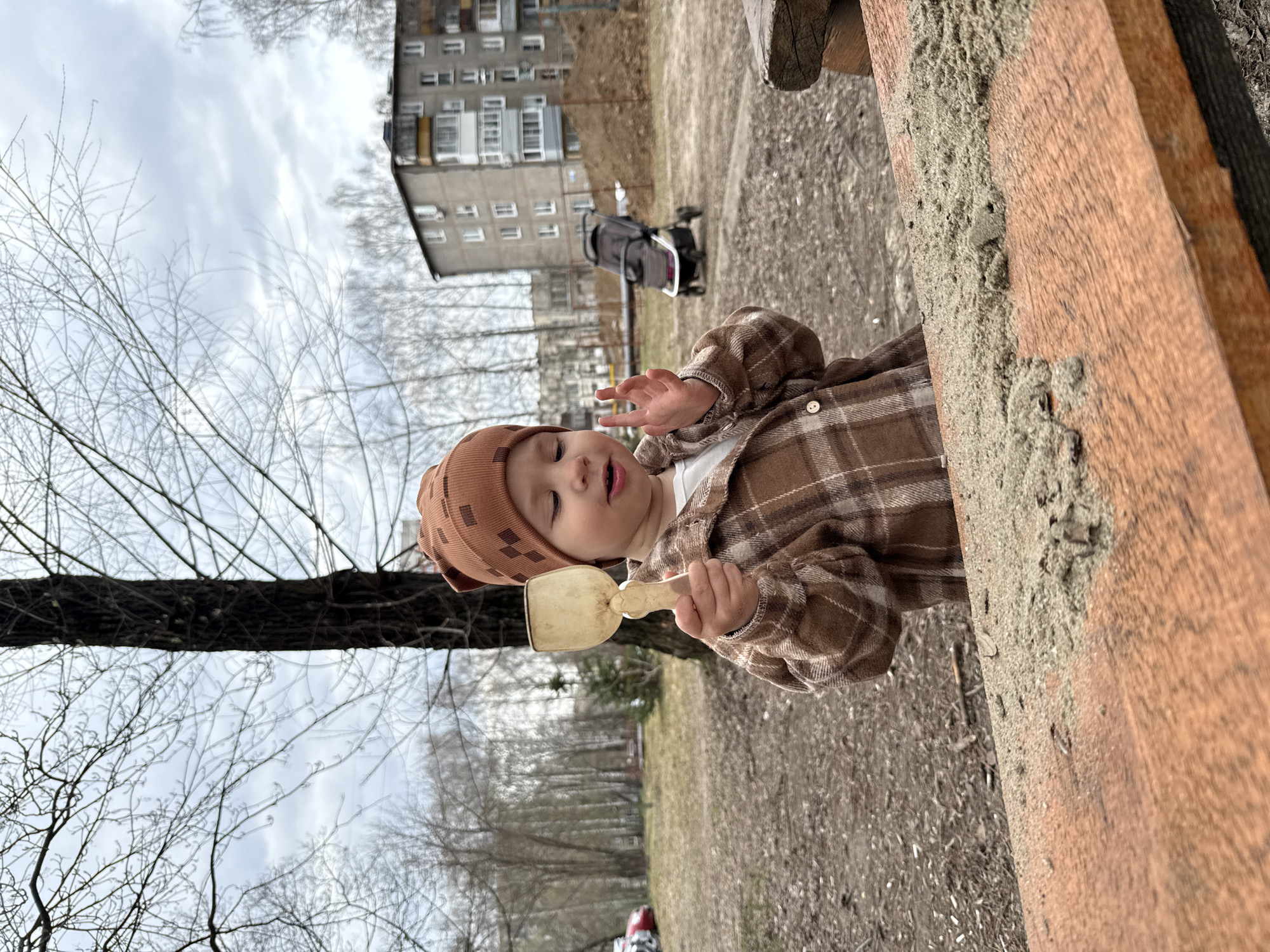 A young toddler is depicted playing outdoors, likely in a sandbox or a similar play area. The child is dressed in warm, practical attire suitable for cooler weather, including a brown plaid shirt, a white undershirt, and a brown beanie hat with a distinctive grid pattern. They are holding a light-colored wooden or plastic shovel, appearing to be focused on manipulating the sand or dirt. The child's eyes are partially closed, and their mouth is slightly ajar, suggesting an absorbed or content expression.
The immediate foreground shows a sturdy wooden border of a play structure, heavily covered with sand or coarse dirt. The ground around the child is a mix of bare soil, sparse dry grass, and debris, characteristic of a natural outdoor setting during a non-growing season.
In the mid-ground and background, the scene reveals an urban or suburban environment. Bare deciduous trees, their branches devoid of leaves, stand against a cloudy, overcast sky, indicating either late autumn, winter, or early spring. A dark-colored stroller is visible further back, hinting at other activities or companions. Dominating the distant background is a multi-story residential building, identifiable by its concrete panel construction and numerous balconies, a common architectural style in Rusanovo, Russia, which matches the provided location context.
The activity taking place is a simple moment of a child's outdoor play. The weather is overcast, with no direct sunlight. The overall atmosphere is quiet and focused on the child's engagement. No visible text is present in the image.