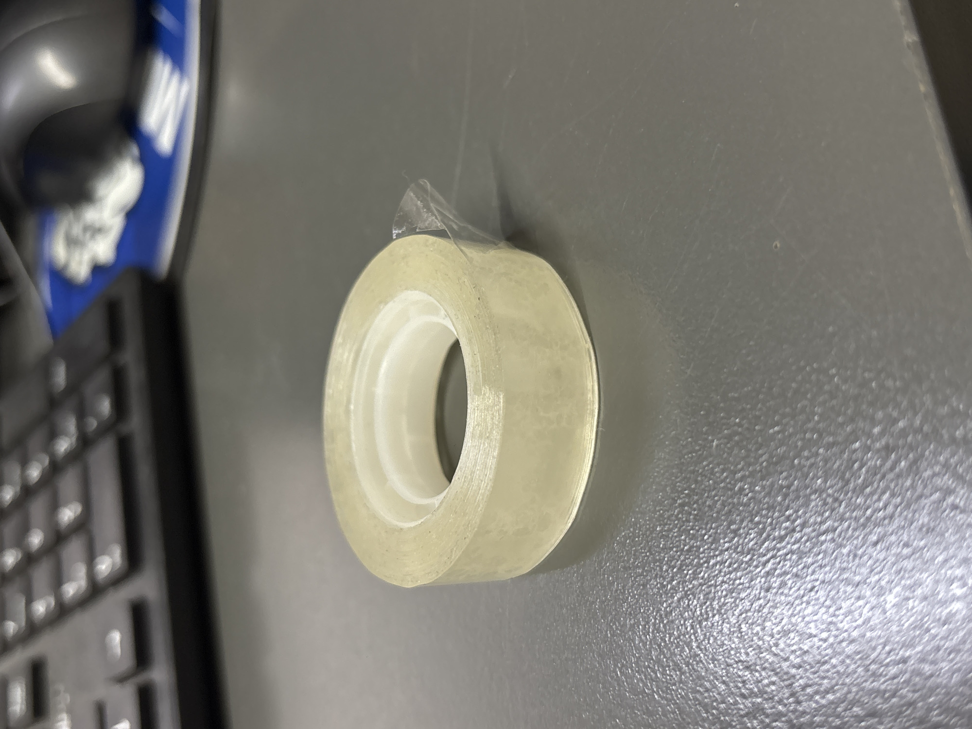 A roll of clear adhesive tape sits on a gray desk. The tape is partially unwound, with a small piece of tape sticking out. In the background, a black keyboard is visible, along with a blurred blue and white object, possibly a mousepad. The scene appears to be indoors, on a desk in an office setting. There are no people or animals present in the image. The lighting is artificial, casting a soft glow on the objects. The image is taken from a slightly elevated angle, looking down at the desk. The overall impression is one of everyday office supplies. There is no information to suggest the location is in Tbilisi, Georgia.